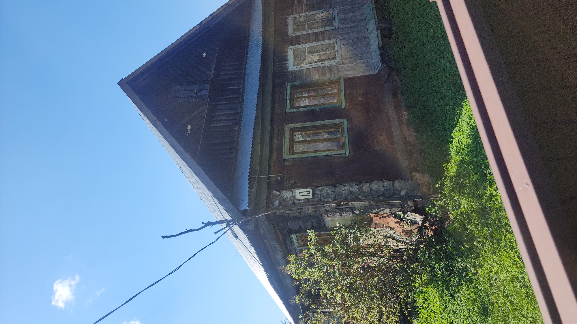 The image depicts a weathered, wooden two-story building with multiple windows, possibly located in a rural or older district. The building's foundation appears to be partially stone, and a number is displayed on the wall. The surrounding environment includes vegetation, suggesting a natural setting. A clear blue sky is visible above the building. There is insufficient information to confirm its location within Moscow, Russia.