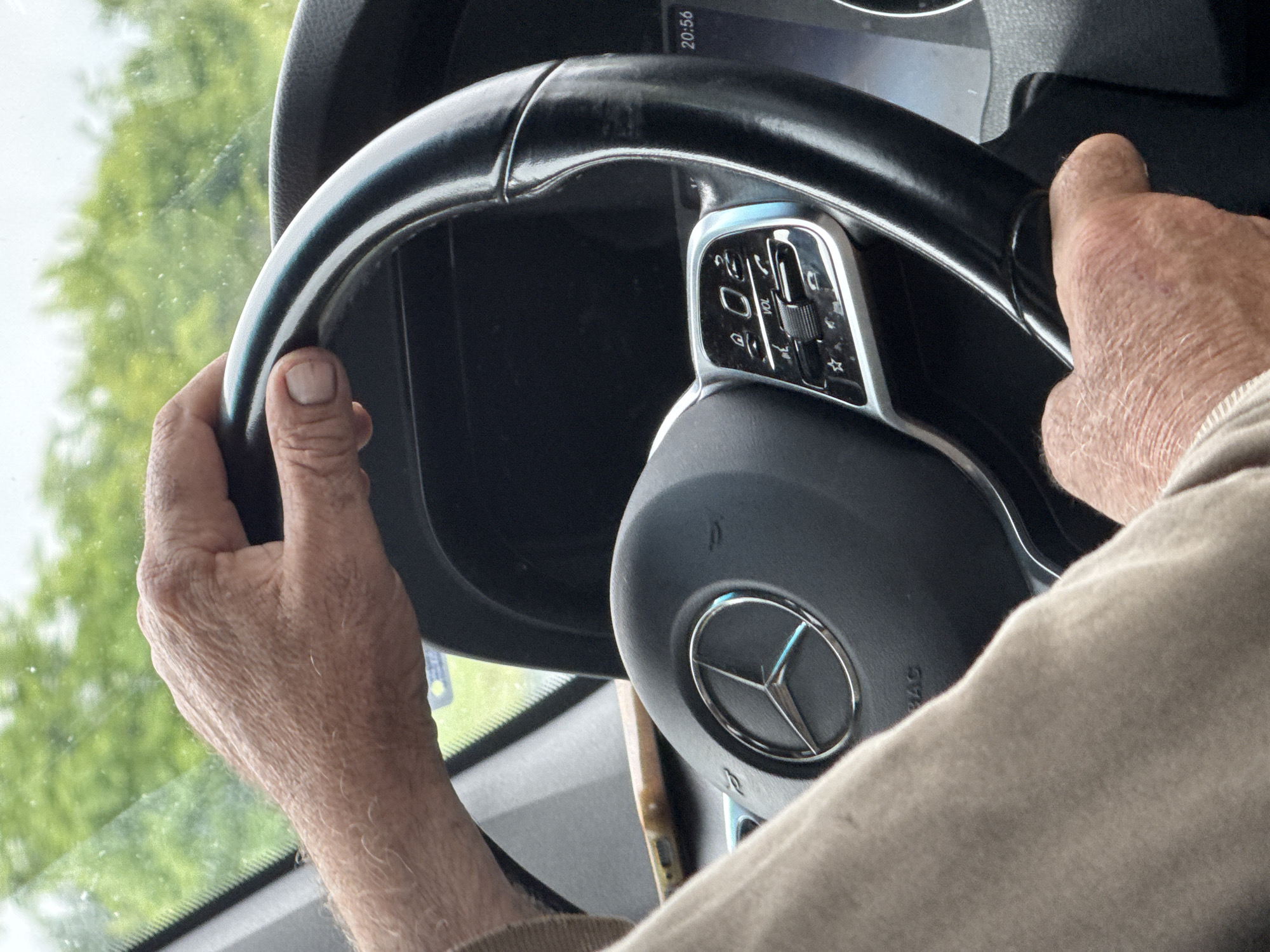 A person is driving a Mercedes-Benz vehicle. The person's hands are visible on the steering wheel. The Mercedes-Benz logo is visible on the steering wheel. The interior of the vehicle is visible. The person is wearing a tan or light-colored shirt or jacket. There is green foliage visible through the windshield. A digital display with the time "20:56" is partially visible in the top right corner. The location is not discernible as being Pleven, Bulgaria, based on the visual information in the image.