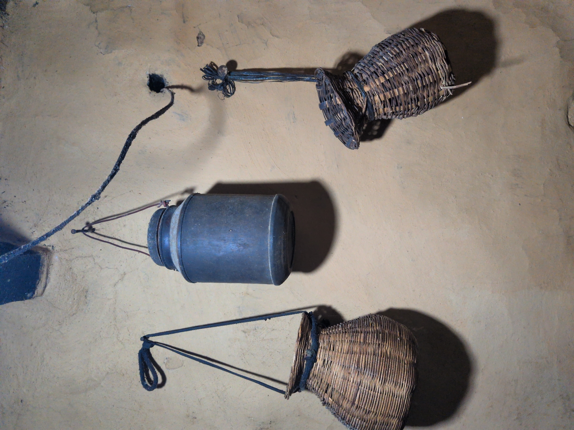A still life photograph taken in Kapurkot, Nepal, depicts two woven baskets and a metal container hanging on a textured, light brown wall. The basket on the left is larger and rounder, suspended by two dark straps. The basket on the right is narrower and more elongated, with a wider opening, hanging by a single braided rope. A dark metal container, cylindrical with a rounded top and bottom, is positioned between the two baskets. The lighting suggests an indoor environment with a shadow cast by each object on the wall. The wall itself appears rough and aged, with some darker markings and cracks visible. A black cord is strung across the upper portion of the image, connecting to a small dark opening in the wall to the right.