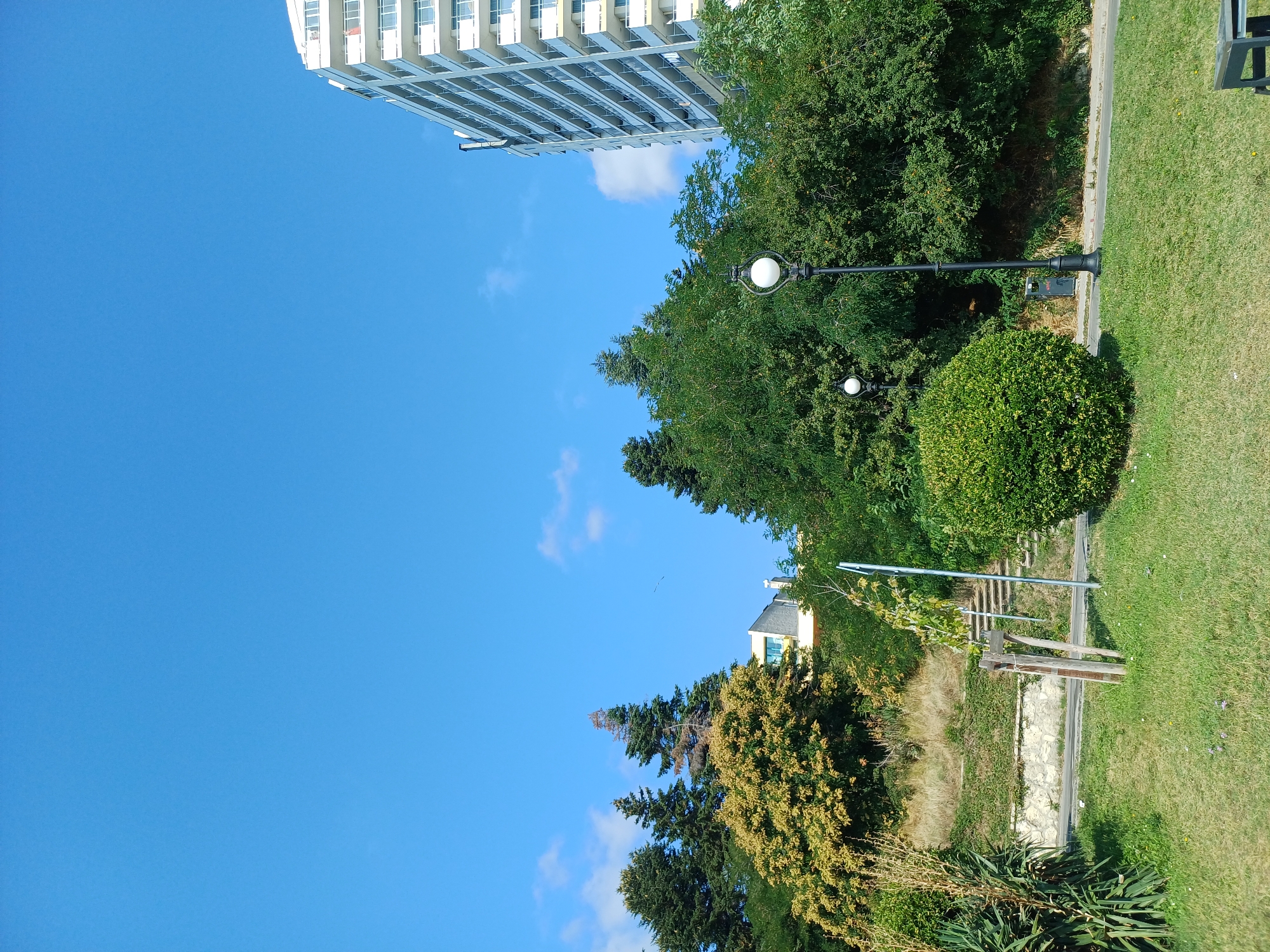 The image shows a scene in Varna, Bulgaria, on a sunny day with a clear blue sky and a few scattered clouds. On the right side of the frame is a tall, white, multi-story building with balconies. In the foreground, there is a well-maintained green lawn. To the left of the lawn, a sloped grassy area is covered with dense green foliage and trees, with a building partially visible behind the trees. Further to the left, there are more trees and vegetation. A black lamppost with a white spherical light fixture stands on the lawn. Stairs are visible ascending a hill in the middle-left background, bordered by a stone wall and covered in green vegetation. A small sapling is staked with wooden poles near the stairs.