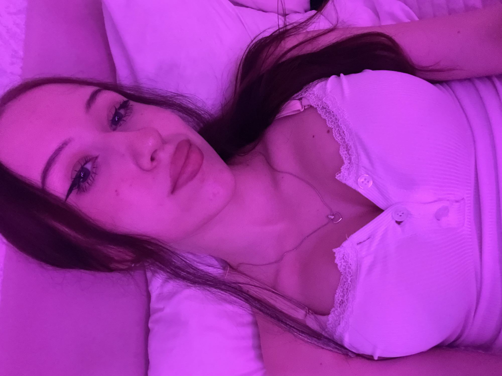 A young woman with long dark hair is lying down, illuminated by pink light. She is wearing a white tank top with lace trim and buttons down the front. A heart-shaped necklace hangs around her neck. Her makeup includes winged eyeliner and dark lipstick. The background appears to be a bed with a white pillow and duvet. The scene is indoors and does not provide any specific details that would allow for identification of a location within Votkinsk, Russia.