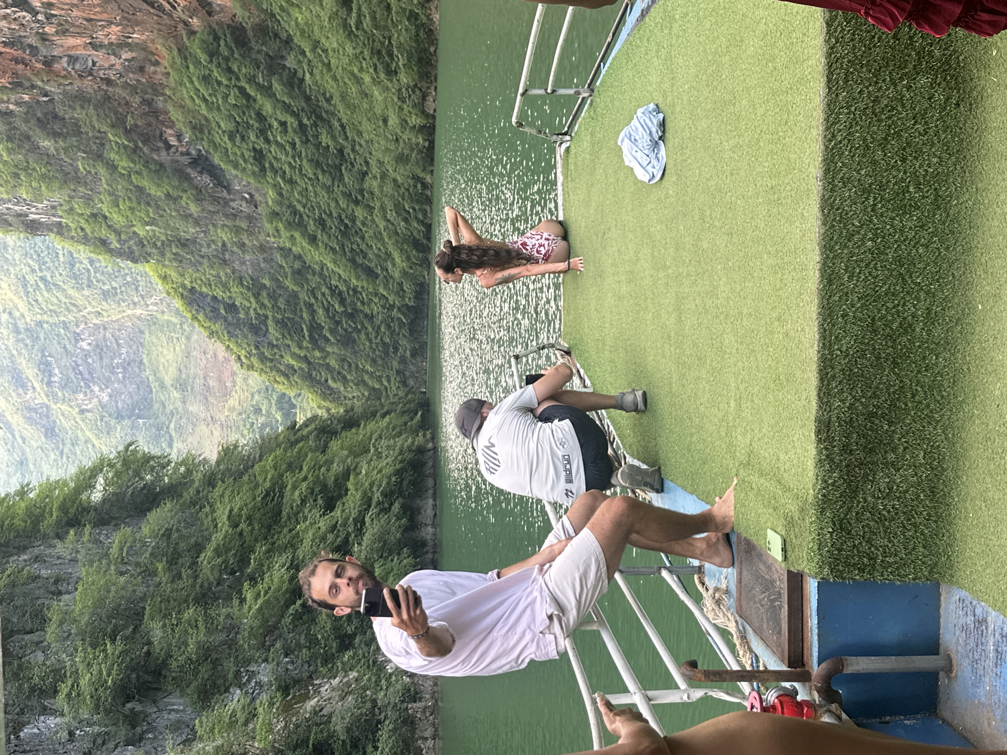 On a boat in Đồng Văn Commune, Vietnam, a man in a white shirt and shorts takes a photo with his phone. Another man in a gray shirt and black shorts kneels near him. A woman in a patterned swimsuit lies on her stomach. The boat has green artificial turf. A cliff covered in green vegetation is visible. The water is green and reflective.