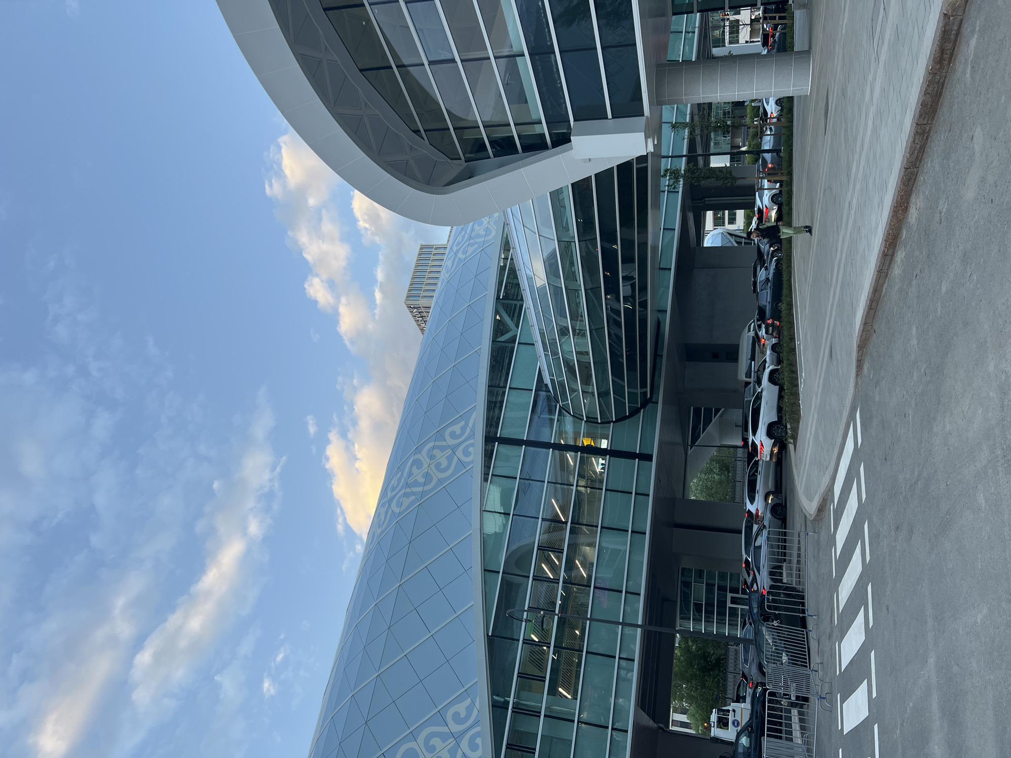 The image depicts a modern architectural structure in Astana, Kazakhstan. The building features a curved façade with glass panels and a decorative blue pattern. The sky is visible with scattered clouds. The foreground shows a pedestrian crosswalk and several parked cars. A person is visible walking in the background.