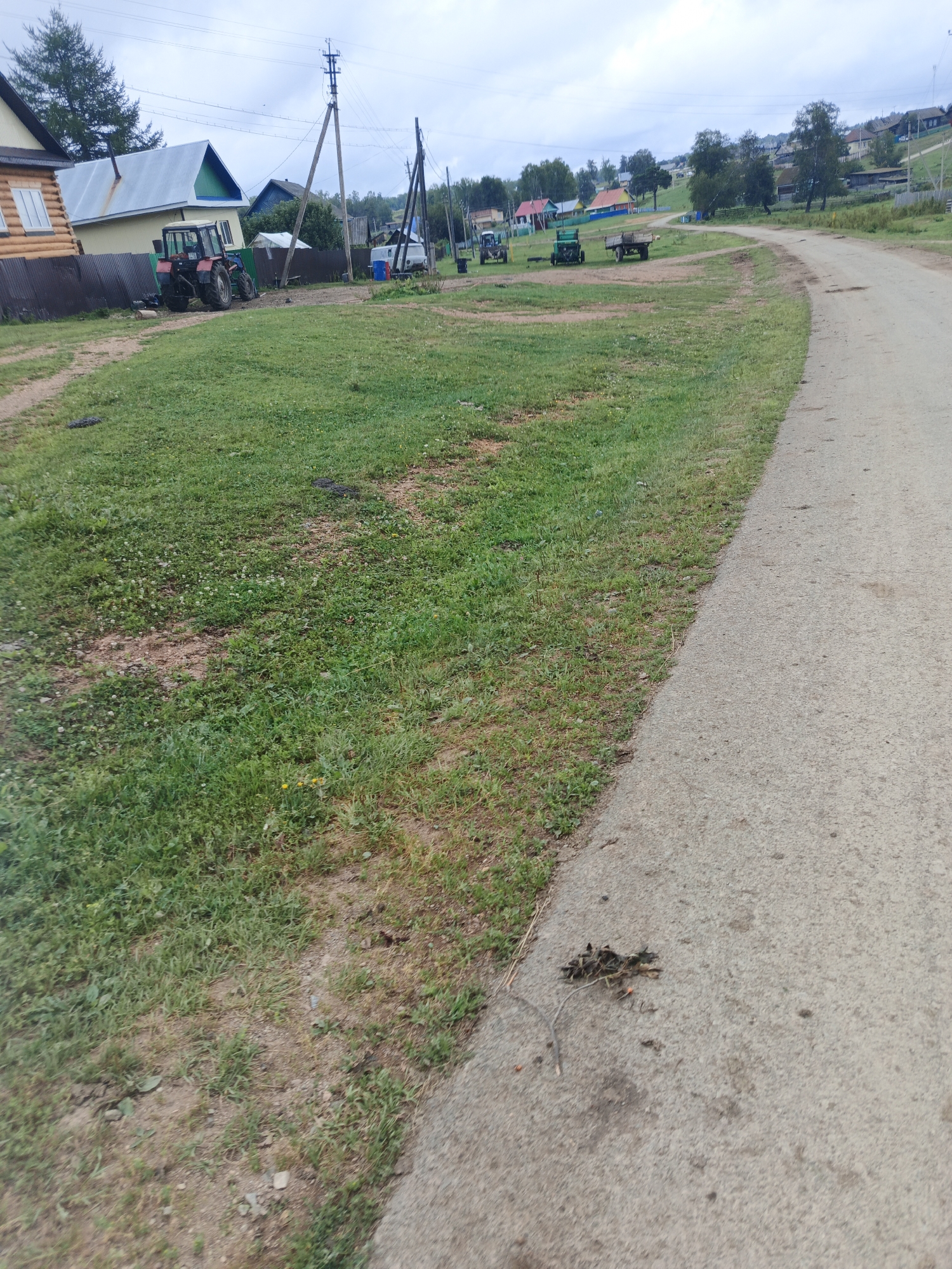 The image shows a rural road in Islambaevo, Russia, with a grassy area and several buildings on the left side. A red tractor is parked in front of a wooden fence, and other vehicles, including a green tractor and a trailer, are visible in the distance. The road is unpaved and curves to the right, leading towards a village with houses and trees. The sky is overcast, and the overall impression is one of a typical rural settlement in Russia.