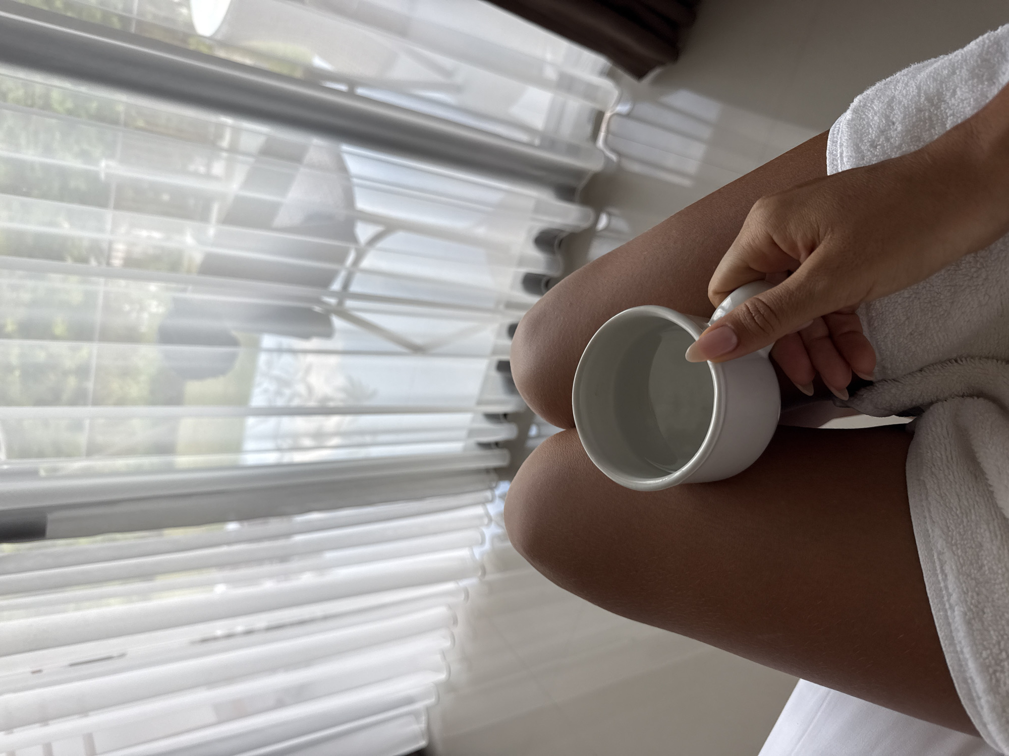 The image displays a first-person, downward perspective of a person's bare, tanned legs, partially covered by a white terrycloth towel or robe. The person's right hand holds a white ceramic mug, filled with a clear liquid, resting on the right leg. The background features an indoor space with a reflective white tiled floor, illuminated by bright natural light. A large window or sliding glass door is covered by sheer, vertically pleated white curtains. Through the translucent curtains, an outdoor scene is visible, characterized by green foliage suggesting trees or bushes. Faintly discernible behind the curtains are elements of a patio or balcony, including a light-colored outdoor chair and a white cylindrical object, possibly a lamp or planter. This modern indoor setting, opening to a tropical-like outdoor view with leisure furniture, is consistent with hotel or resort accommodations typical of tourist areas such as Kuta Selatan, Indonesia.
