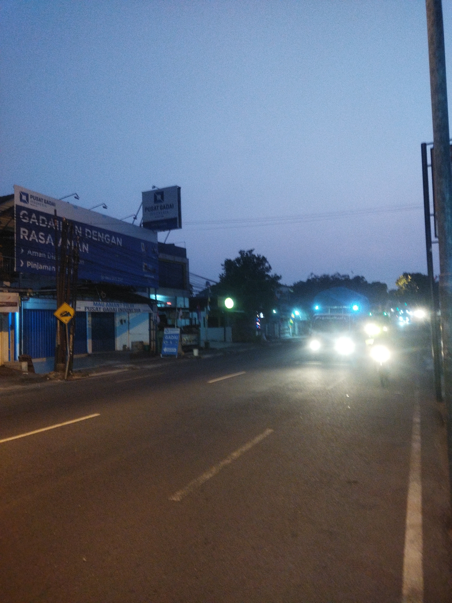 An outdoor medium shot captures a street scene in the city of Wonogiri, Indonesia, during what appears to be dusk or early evening. The sky is a pale, hazy blue. On the left side of the image, a building displays a large blue banner with white text that reads "PUSAT GADAI INDONESIA" and "GADAIAN DENGAN RASA AMAN". Below this, smaller text lists "Aman Cie" and "Pinjaman". Adjacent to this banner, a yellow diamond-shaped sign with a black symbol is mounted on a pole. Further down the street, another building has a sign that reads "MAU AMAN FIUR PUSAT GADAI INDONESIA". On the right side of the road, several vehicles are visible with their headlights on, illuminating the asphalt. A truck with bright blue lights on its roof is prominent in the center of the road. The road itself is dark asphalt with faint white lane markings. A tall, dark pole stands on the far right edge of the frame, partially obscuring the background. Trees and other structures are dimly visible in the distance. The overall atmosphere is that of a typical Indonesian street at twilight, with commercial signage and traffic contributing to the urban environment.