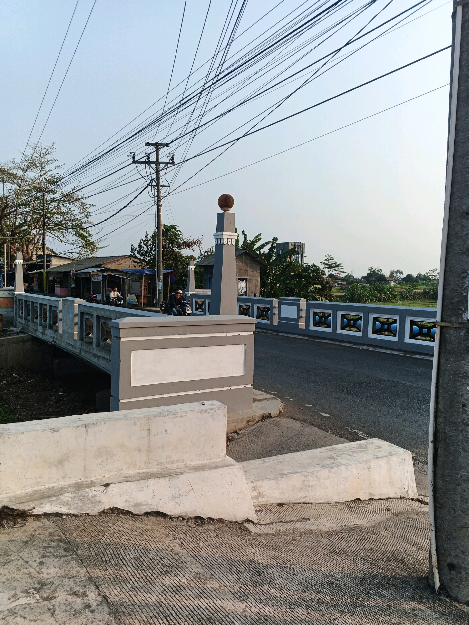 The image depicts a bridge spanning a small body of water, likely a canal or ditch, in a semi-urban or rural area of Cilacap, Indonesia. The bridge is constructed of concrete with grey and white decorative elements along its railings, featuring repeating square patterns. A paved road runs across the bridge and continues to the right. On the left side of the bridge, a few small buildings with tin roofs and what appear to be market stalls are visible, with a few people on motorcycles. Overhead, a dense network of utility wires is strung between poles, creating a cluttered sky. The environment includes some trees and vegetation, indicating it is not a dense urban setting. The foreground shows a concrete embankment with some cracks and a textured surface. A large, grey utility pole stands prominently on the right side of the image. The scene appears to be captured during daylight, with the sun casting soft shadows.