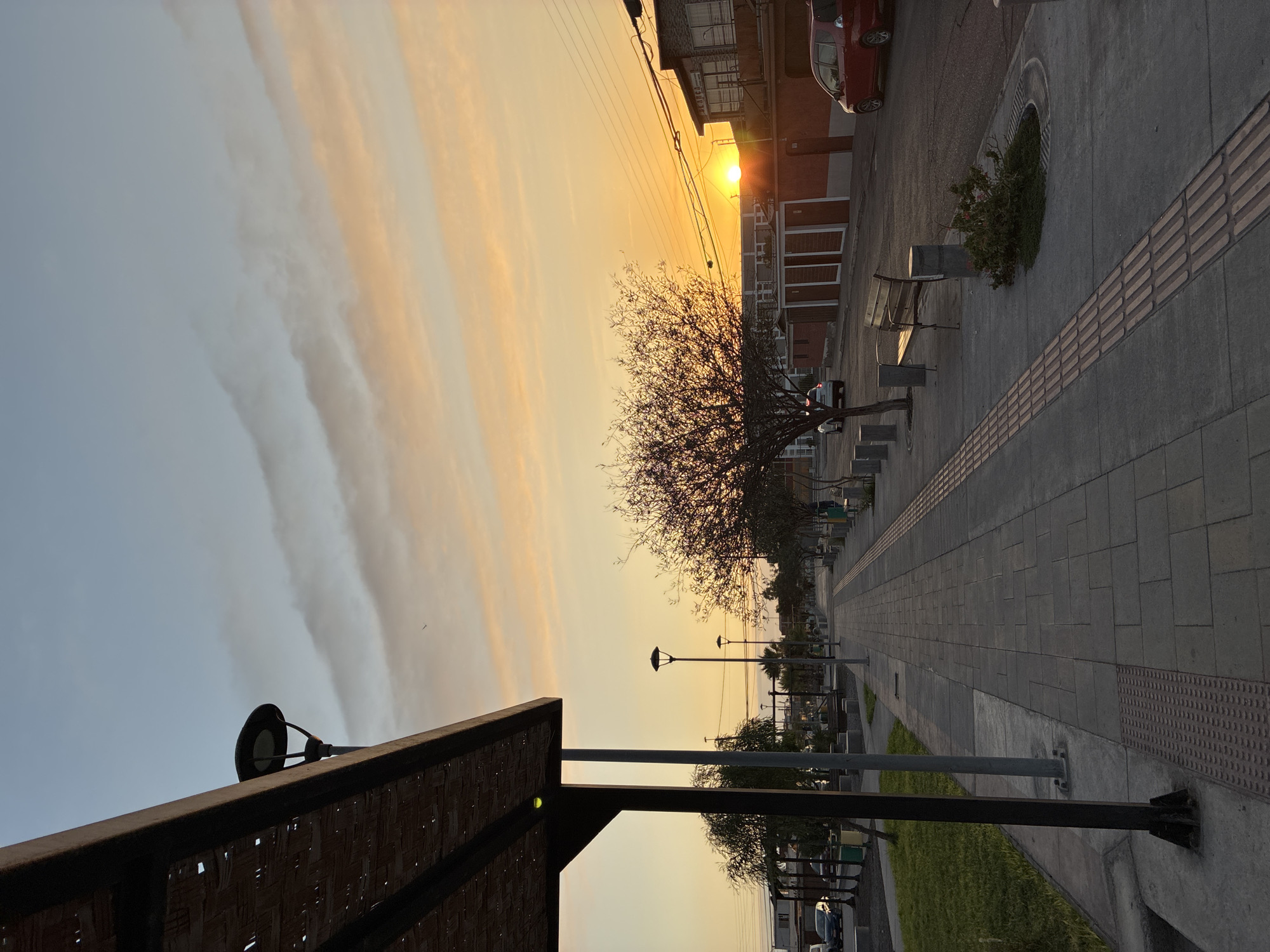 The image shows a street scene in Arica, Chile during a sunset. The sky is filled with streaks of orange and gray clouds. A bare tree stands along the right side of the walkway. A paved path runs along the right side, with benches, lampposts, and small patches of greenery lining the street. In the distance, the ocean is visible. On the far right of the image, there is a building with a red car parked near it. The sun is shining through the building. In the foreground, there is a partial view of a structure with a dark, patterned roof.
