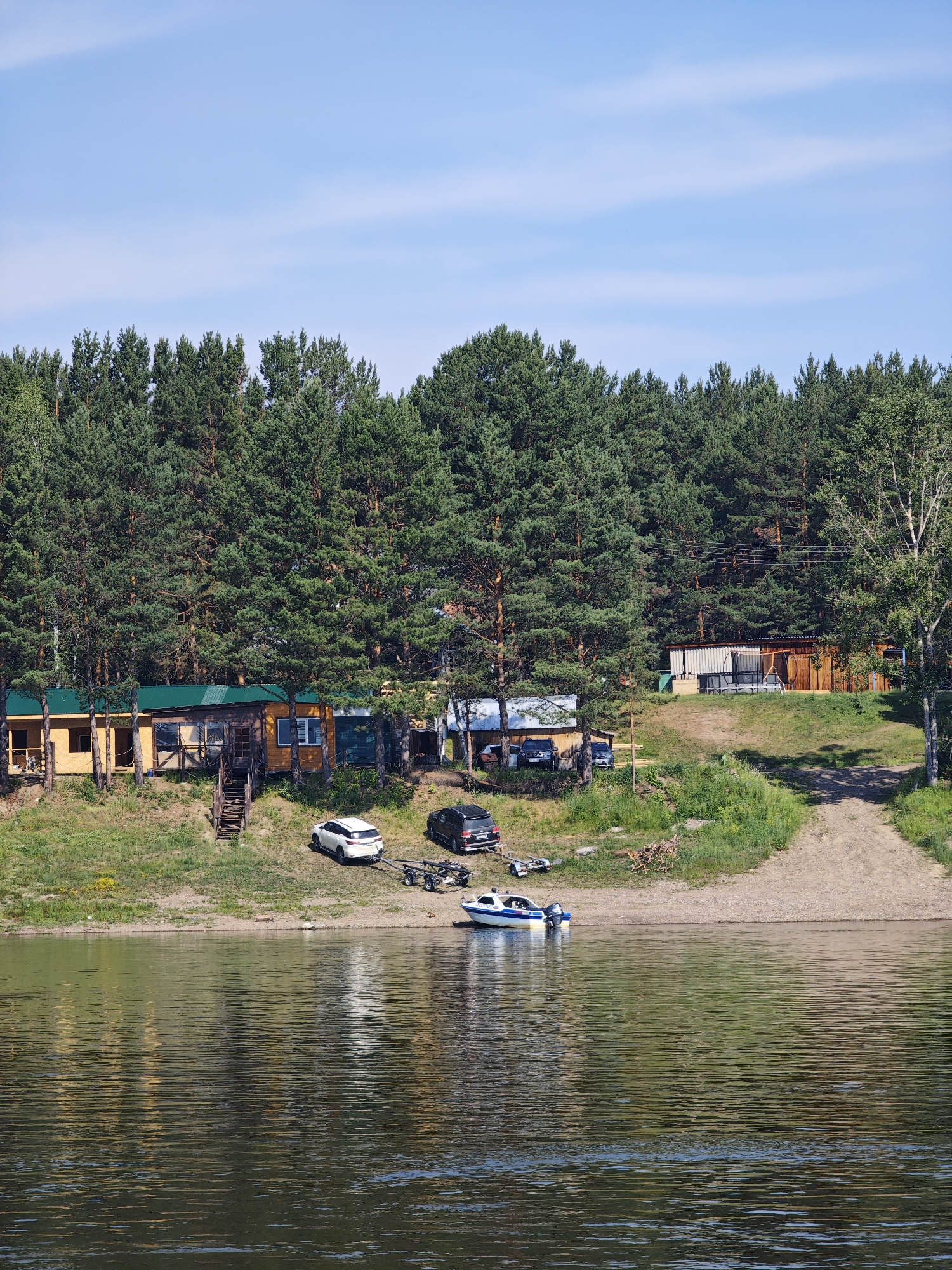The image depicts a serene waterscape with a forested background and a small settlement along the bank. A wide, calm river occupies the foreground, its surface rippling and reflecting the green of the trees and the blue of the sky. The far bank is lined with a dense forest of tall pine trees.
Nestled at the base of the forest, on a slightly elevated grassy embankment, are several wooden structures, likely small houses or cabins, with green roofs. In front of these structures, a sandy area slopes down to the water. Several vehicles are parked on the embankment, including a white SUV towing a boat trailer and a black SUV. A small motorboat is docked near the shore. A wooden staircase leads up from the sandy area to one of the buildings. The sky above is a clear, bright blue with a few wispy clouds. The overall impression is one of a peaceful, rural setting, possibly a recreational area or a small village on the outskirts of Podporozh'e, Russia, given the context of the query.