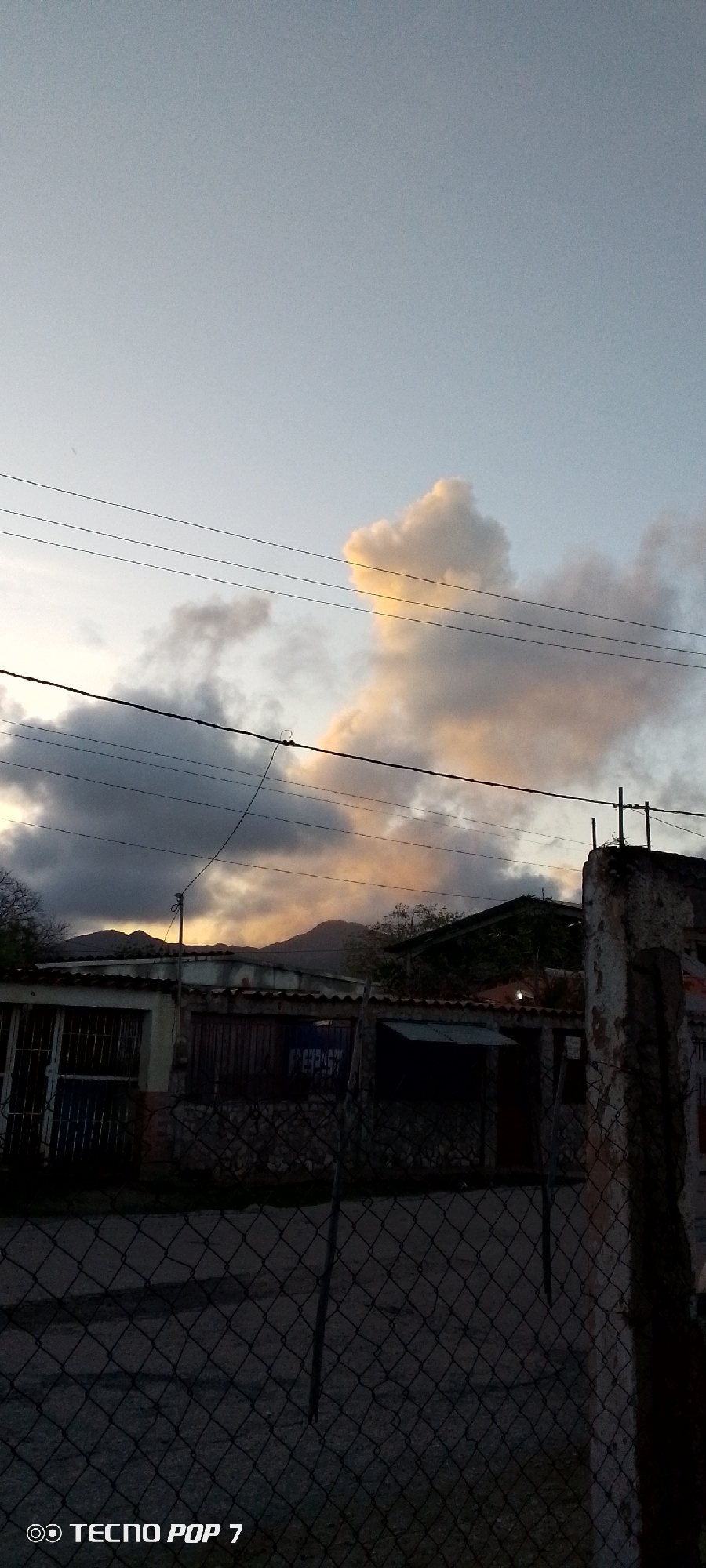A photograph taken at dusk in Altagracia, Venezuela, depicts a street scene dominated by a partially overcast sky transitioning from day to night. The sky features dramatic clouds, some illuminated with warm, golden hues from the setting sun, while others appear dark and heavy. Power lines crisscross the upper portion of the frame.
In the middle ground, a row of low-rise buildings lines the street, some with metal gates and tiled roofs. One building has a sign with partially visible text that may read "PEPSI". A chain-link fence runs along the foreground, obscuring parts of the street and the buildings. The overall impression is of a residential or commercial area in a state of twilight, with a focus on the atmospheric sky and the urban infrastructure. The bottom of the image includes a watermark "TECNO POP 7".