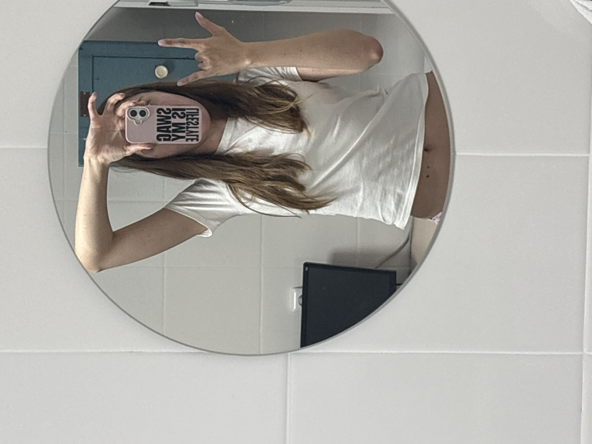 A person is captured in a mirror selfie, with the image rotated 90 degrees counter-clockwise. The individual has long brown hair and is wearing a white t-shirt. Their face is obscured by a light pink smartphone, which has a black and white sticker on its back that reads "SWAG IS MY LIFESTYLE" (visible as a mirror reflection). The person's left hand (appearing on the right side of the reflection) is raised, forming a "rock on" gesture.
The round or oval-shaped mirror reflects the person's upper body. In the background of the reflection, a dark teal or blue cabinet with a white circular knob is visible. A small portion of the person's midriff is exposed below the t-shirt, showing their navel and two small marks, possibly moles or piercings. A sliver of light pink or purple undergarment is also discernible. Outside the mirror's frame, the wall is covered in white tiles with visible grout lines, suggesting an indoor setting, likely a bathroom. A black rectangular object, possibly an electronic device, is partially reflected in the lower right section of the mirror.