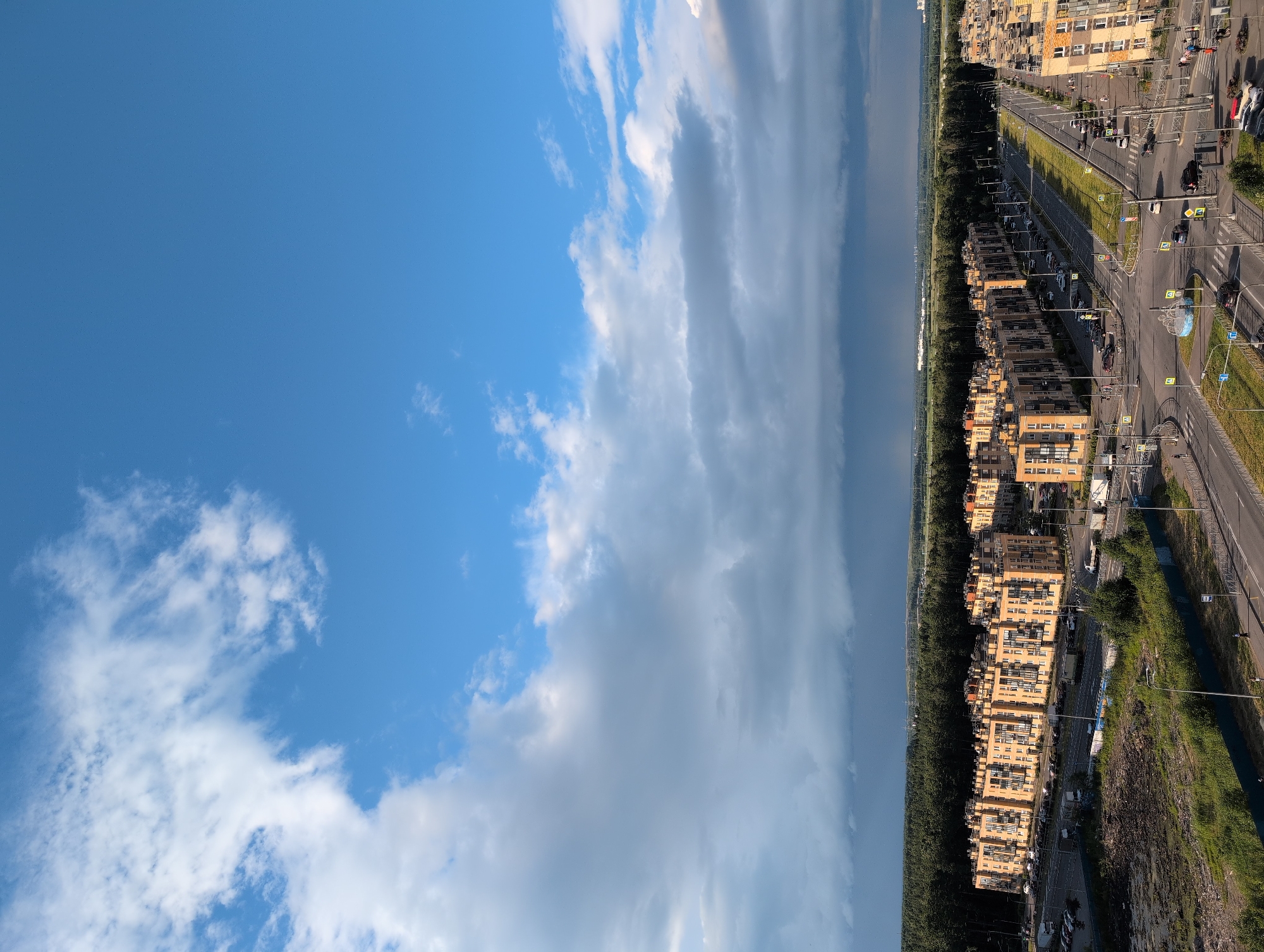 The image shows an aerial view of a residential area in Saint Petersburg, Russia. The scene is dominated by a bright blue sky with scattered white clouds on the left, transitioning to darker, more dense clouds towards the right. Below the sky, a long line of modern, multi-story apartment buildings stretches horizontally across the frame. These buildings are primarily light-colored with many windows.
Adjacent to the buildings is a wide, paved road with traffic. Several cars are visible on the road, along with streetlights and traffic signals. There are also sidewalks and grassy areas bordering the road and buildings. A dense line of dark green trees runs alongside the apartment complexes, separating them from what appears to be a body of water or a long, flat expanse in the distance on the far right. The time of day appears to be daytime, likely late morning or afternoon, given the bright sunlight casting shadows. The weather seems to be dynamic, with clear skies on one side and potentially stormy clouds on the other. The location context provided suggests this is a suburban or outer-city area of Saint Petersburg. No visible text is present in the image. The overall impression is of a modern, urban landscape under a vast sky.