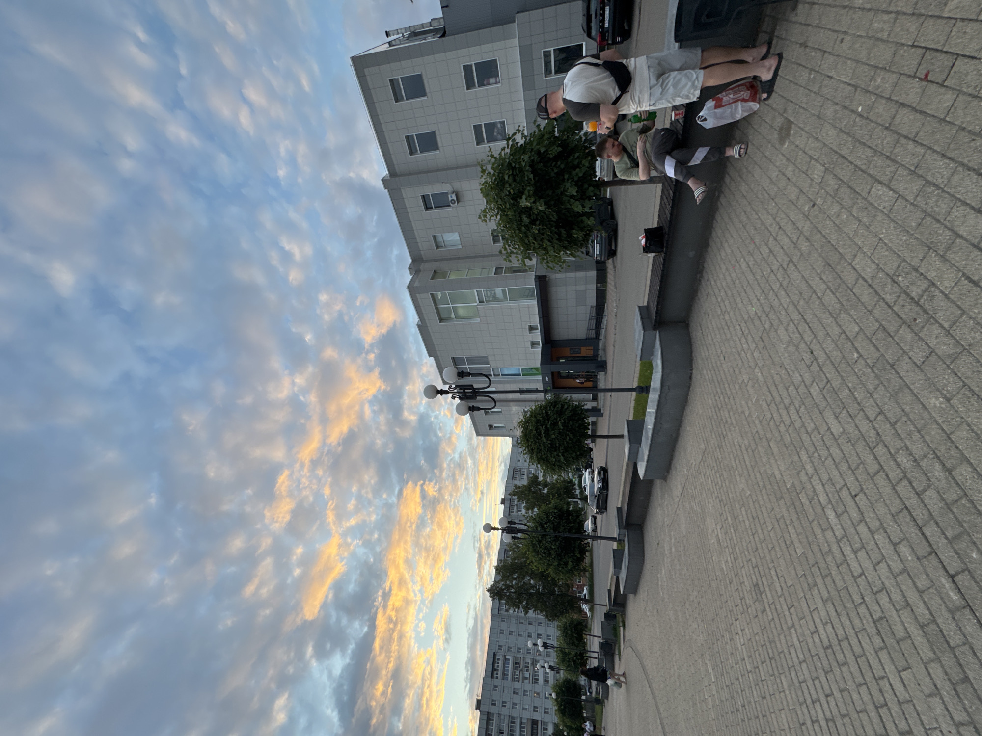 The image depicts an outdoor scene in the city of Chaikovsky, Russia. In the foreground, a paved area with brickwork leads towards a street lined with trees and modern buildings. The sky is partially cloudy with hues of orange and yellow from what appears to be a sunset or sunrise.
On the right side of the image, two men are visible. One is sitting on a bench, wearing a green t-shirt and dark patterned pants, and holding a green bottle. The other man is standing, wearing a light-colored t-shirt, dark shorts, and a backpack, and holding a beverage with an orange liquid. A white plastic bag with red print is on the ground near the standing man.
In the middle ground, there are several streetlights and neatly trimmed trees. Cars are parked and driving on the road behind the trees. To the left, a tall, light-colored residential building is prominent. The overall environment suggests a public space within a city, likely a park or a pedestrian area adjacent to a street.