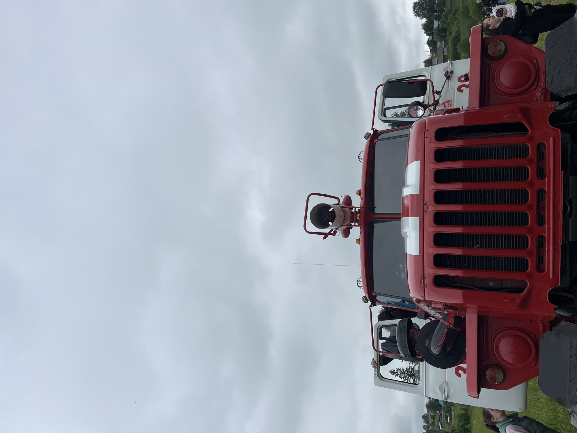 The image presents a low-angle, head-on view of a large, red and white fire truck situated outdoors under a heavily overcast, grey sky in the city of Unknown, Unknown. The truck's prominent red cab features a vertical black grille, a horizontal white stripe across the top of the grille, and large round headlights with smaller round signal lights beneath them. A large, elevated water monitor or cannon is mounted centrally on the roof, flanked by two small amber warning lights. A thin whip antenna extends vertically from the right side of the cab roof.
Both the driver's side (left) and passenger's side (right) cab doors are open. The driver's side door is white and reveals a large, black air intake/filter assembly mounted externally to the cab. A person's hand is visible grasping the frame of the open driver's side door. The passenger's side door is also white, with faint red Cyrillic text and the number "20" visible on the white side panel behind it.
On the left side of the foreground, a woman wearing a light green vest over a light top leans forward, her head positioned near the truck's front fender, seemingly looking into the open driver's side or at something low on the truck. On the right side, a second woman dressed in dark clothing holds a small child clad in a light-colored, possibly hooded, outfit. Both the woman and child are looking towards the right, away from the immediate front of the truck.
The background consists of rolling green hills dotted with multiple residential buildings of a rural settlement and scattered coniferous trees. The immediate foreground is covered in sparse green grass. The overall lighting is soft and diffuse, consistent with an overcast day.