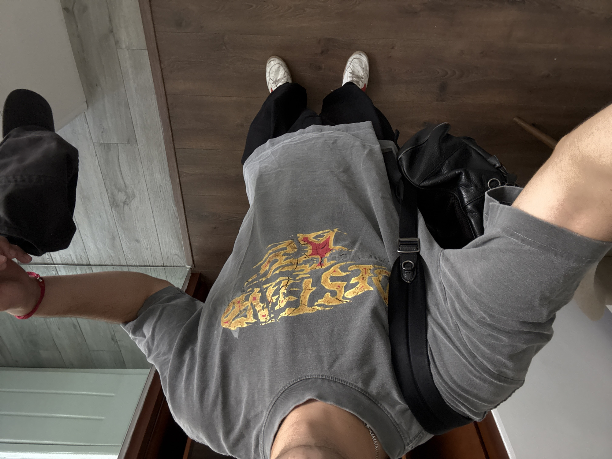 A first-person perspective photograph shows a person standing indoors on a wood-look floor. The person is wearing white sneakers, black pants, and a gray t-shirt with a gold graphic. A black bag hangs off their shoulder. Part of a gray chair is visible on the left and a white wall on the right.
This image cannot be located in San Bernardino Tlaxcalancingo, Mexico, without more specific information.