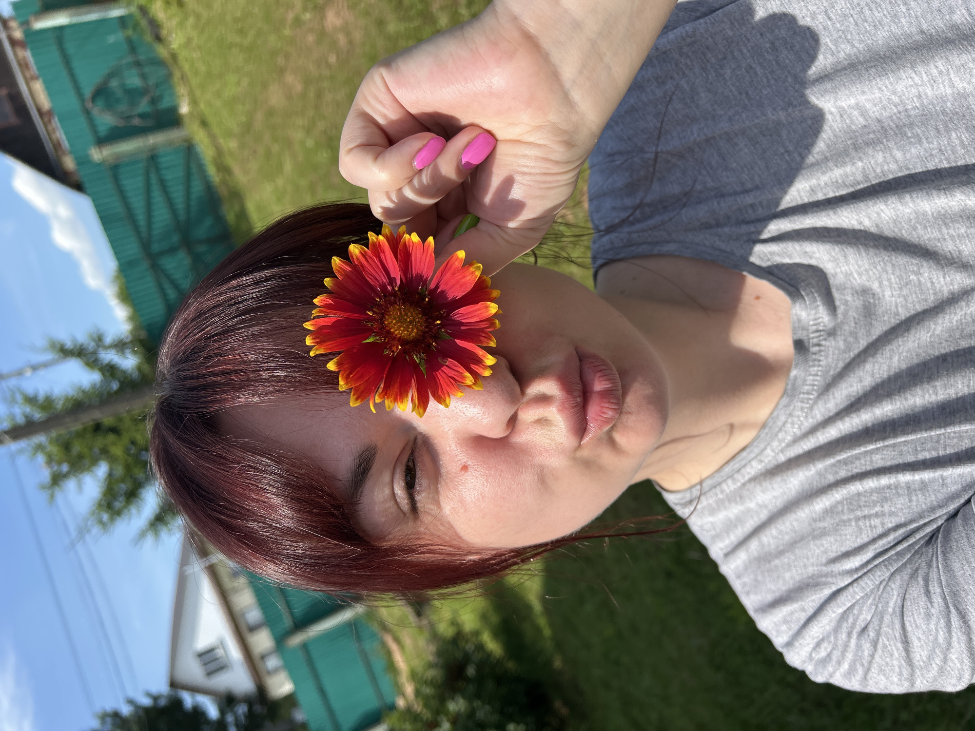 In a close-up, outdoor selfie, a woman with auburn hair and a grey t-shirt is holding a red and yellow flower in front of her right eye. She is puckering her lips and her left eye is visible. Her right hand, with bright pink nail polish, is holding the flower. The background consists of a green fence, a grassy area, trees, a house, and a blue sky with some clouds, suggesting a rural or suburban setting likely in the city of Фоки, Russia.