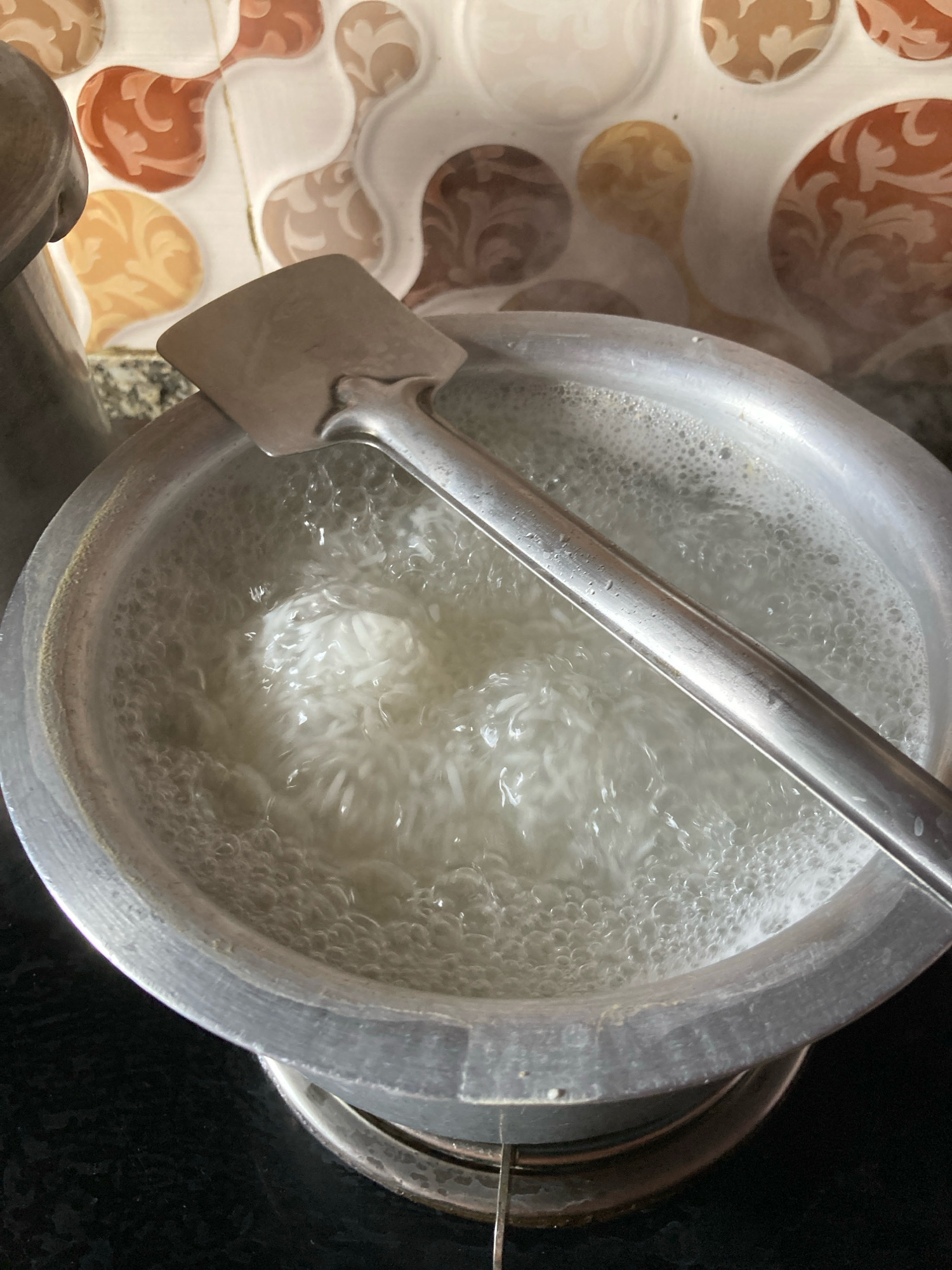 The image shows a close-up view of a metal pot filled with boiling water and some white, stringy food item, likely noodles or rice, being cooked. A metal spatula rests across the rim of the pot, partially submerged in the bubbling water. The pot is situated on a black stovetop. In the background, there is a tiled wall with an abstract, organic pattern in shades of brown, orange, and beige. The lighting suggests an indoor setting, possibly a kitchen. The image does not provide any specific visual cues that would allow for the determination of its location in Kolkata, India.