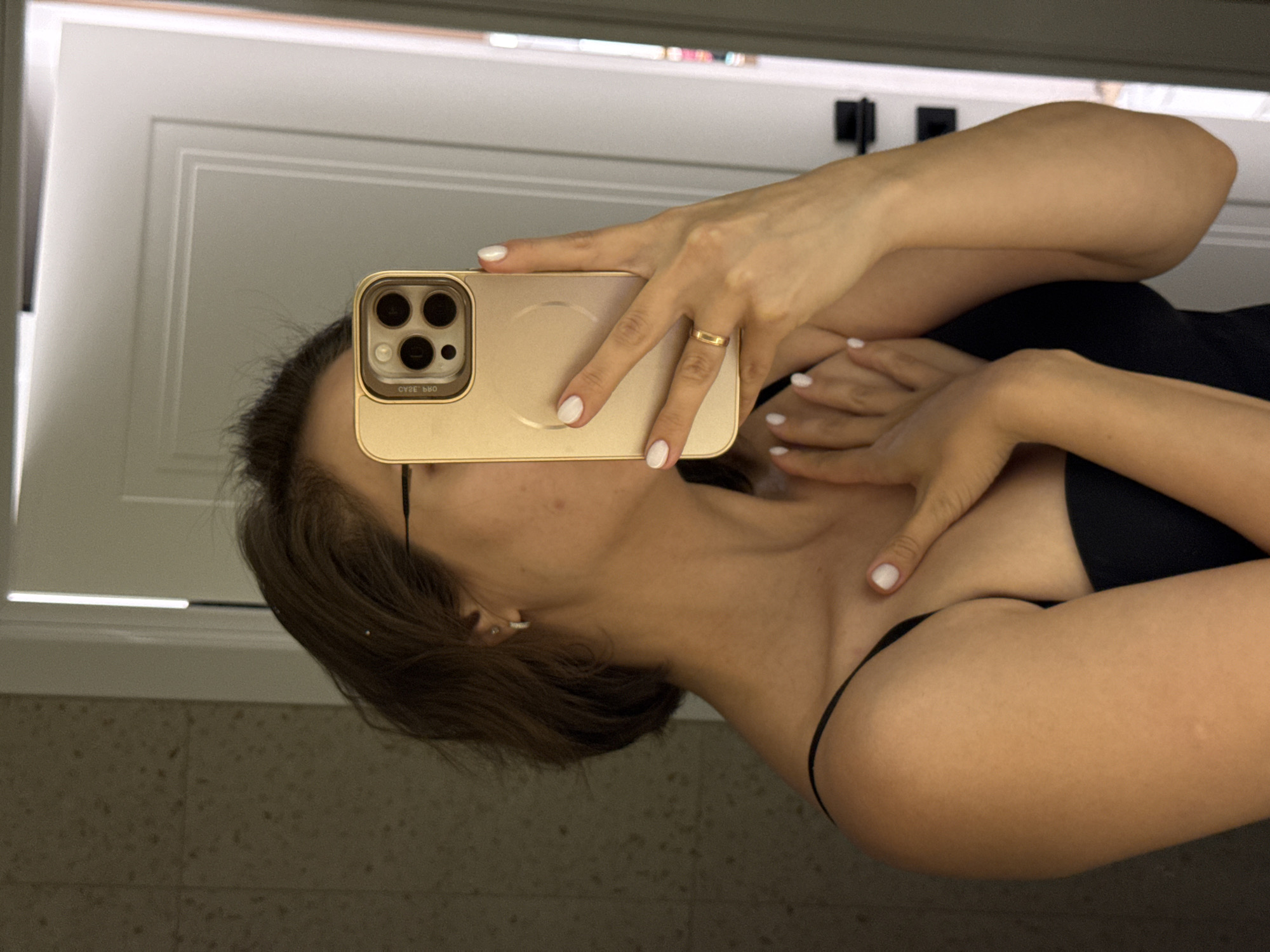 A woman with brown hair is taking a selfie in a bathroom with a gold iPhone. She is wearing glasses and a black top. Her fingernails are painted white, and she is wearing a gold ring. The background includes a white cabinet and a beige tiled wall. There is no information suggesting the photo was taken in Pervomaysky, Russia.