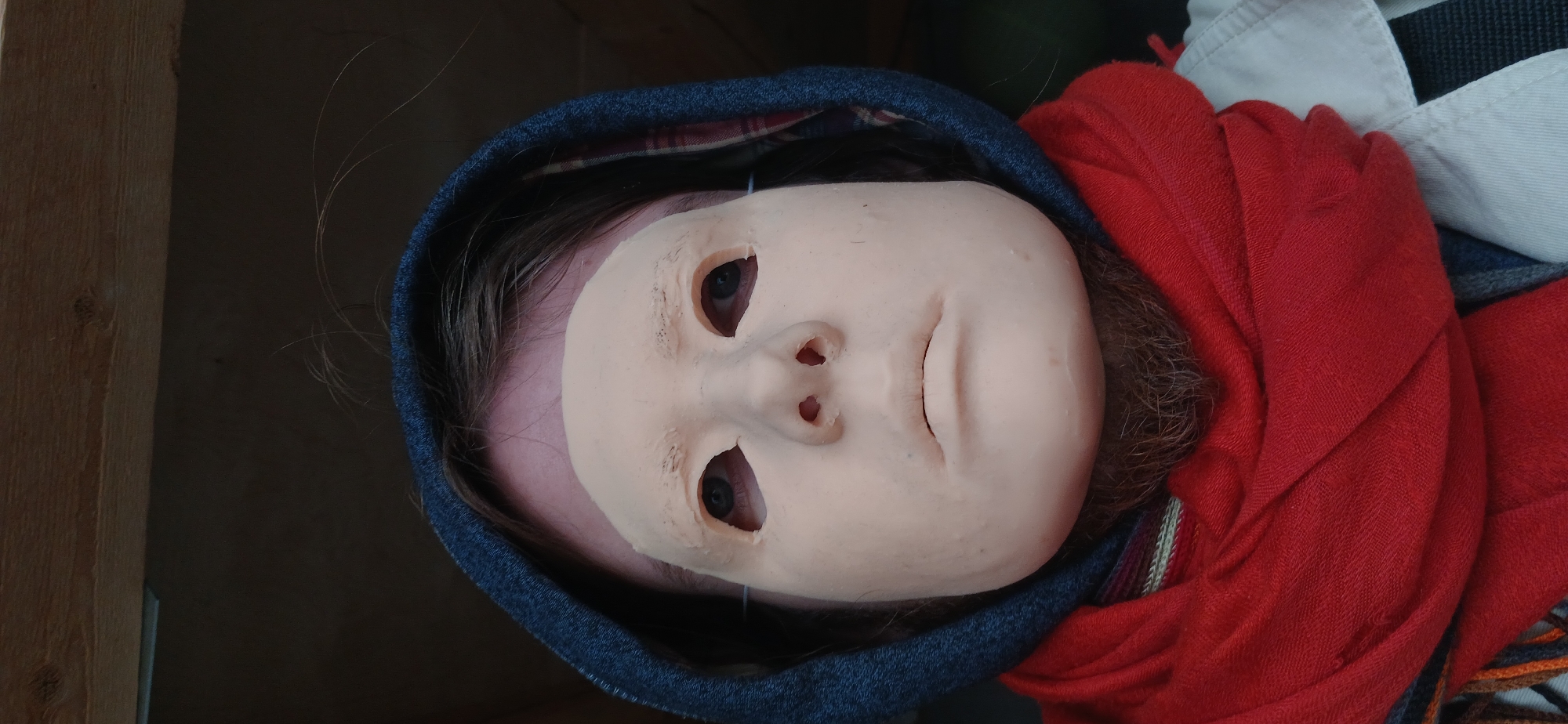 A close-up portrait shows a person wearing a pale, mask-like face covering. The smooth, light-colored mask features cutouts for the eyes, revealing dark pupils, and small openings for the nostrils. The mouth area of the mask is molded shut. Dark hair is visible around the edges of the mask and beneath a dark blue hooded garment, which is worn with the hood up. A vibrant red, textured scarf is wrapped around the person's neck and shoulders, contrasting with the dark blue hood and the pale mask. Below the scarf, a white collar with blue stripes is partially visible. The background on the left consists of a light brown wooden beam or plank, while the rest of the background is dark and indistinct. The lighting is soft. No text is visible.