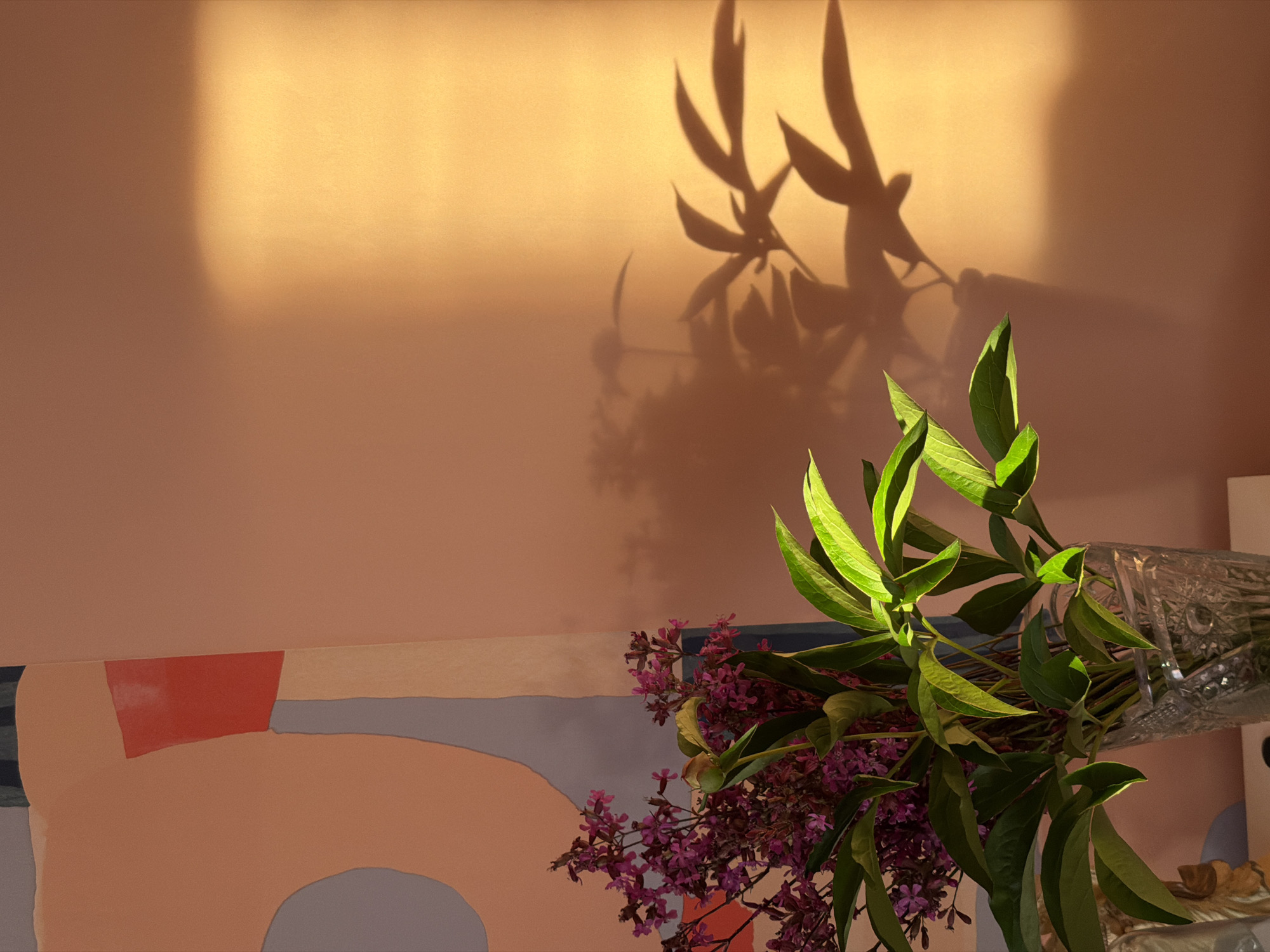 This is a close-up, indoor, still-life photograph of a bouquet of purple flowers and green leaves in a clear glass vase. The bouquet is positioned in the foreground, with the flowers and leaves partially obscuring the vase. Behind the flowers is a large, abstract painting on an easel, with sections of peach, grey, and red. The background is a peach-colored wall, bathed in warm, golden light that casts shadows from the foliage onto the wall. The photograph appears to have been taken in Chaikovsky, Russia, given the prompt's instructions. There are no people or actions depicted in the image.