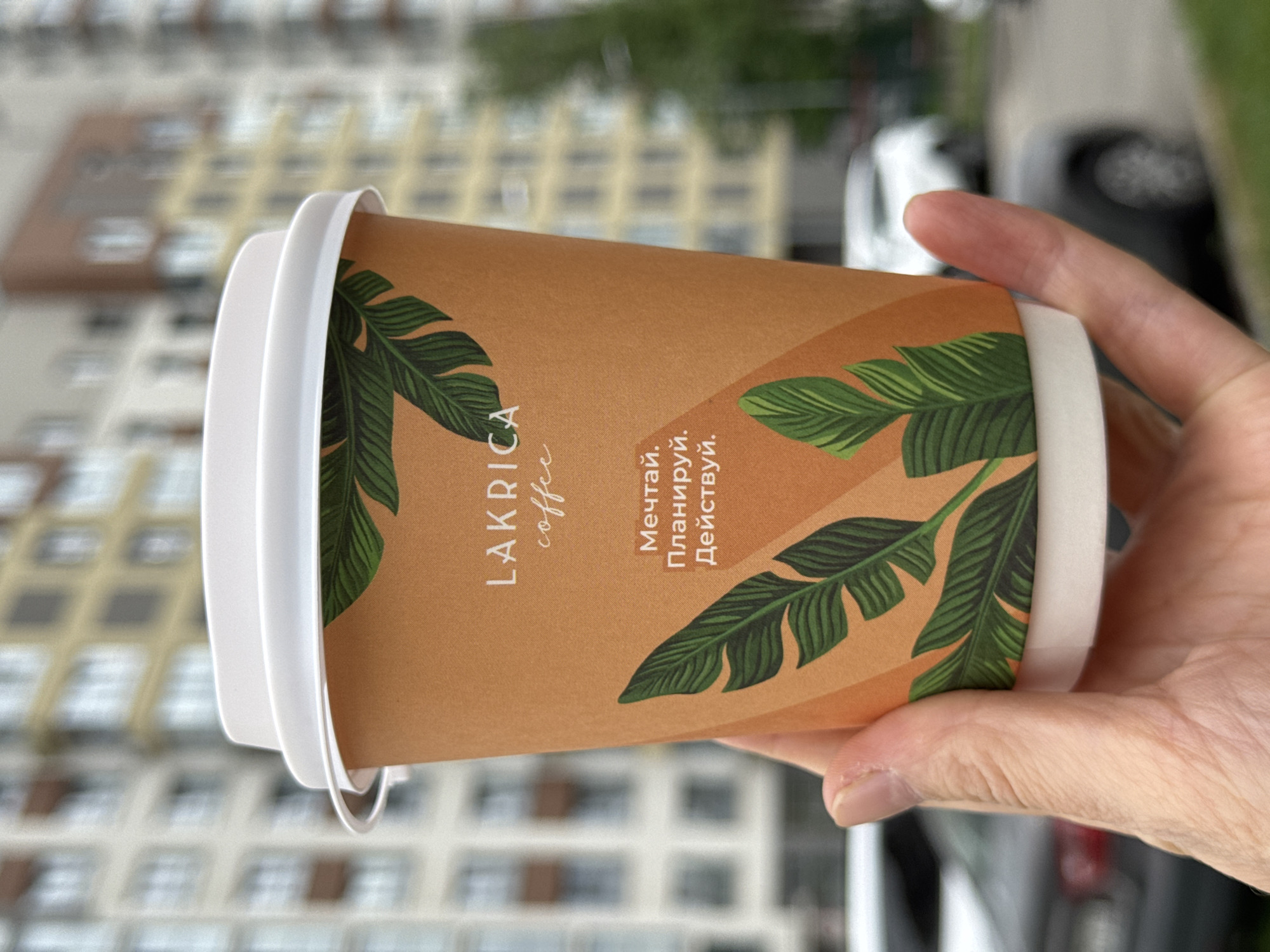 A hand holds a tan paper coffee cup with a white lid. The cup has a leafy design and the words "LAKRICA coffee" printed on it, along with text in Russian that translates to "Dream. Plan. Act." The background shows an out-of-focus apartment building, indicating an urban setting. Based on the "LAKRICA coffee" brand and apartment architecture, the location is likely Izhevsk, Russia. Parts of a white vehicle and a black vehicle are partially visible and out of focus in the background.