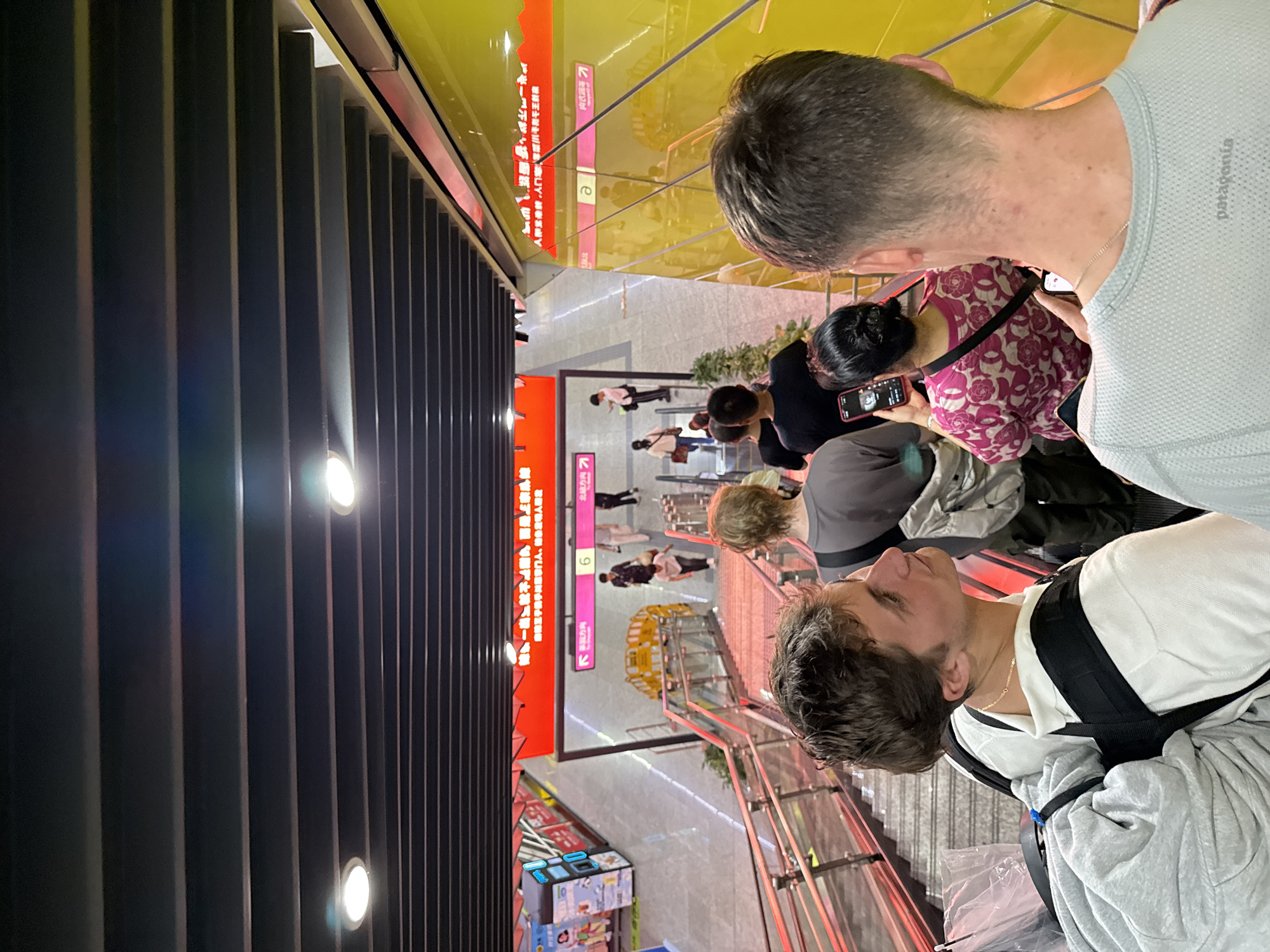 The image captures a scene inside a transportation hub, possibly a subway station, within Yuzhong District, China. Several individuals are present, focused on their mobile devices, waiting, and traveling via escalators. Signage in Mandarin Chinese indicates station information. Bright lights illuminate the area. Storefronts displaying retail items, potentially vending machines, can be seen along one side. A barrier made of vertical posts is along one side of the escalators.
