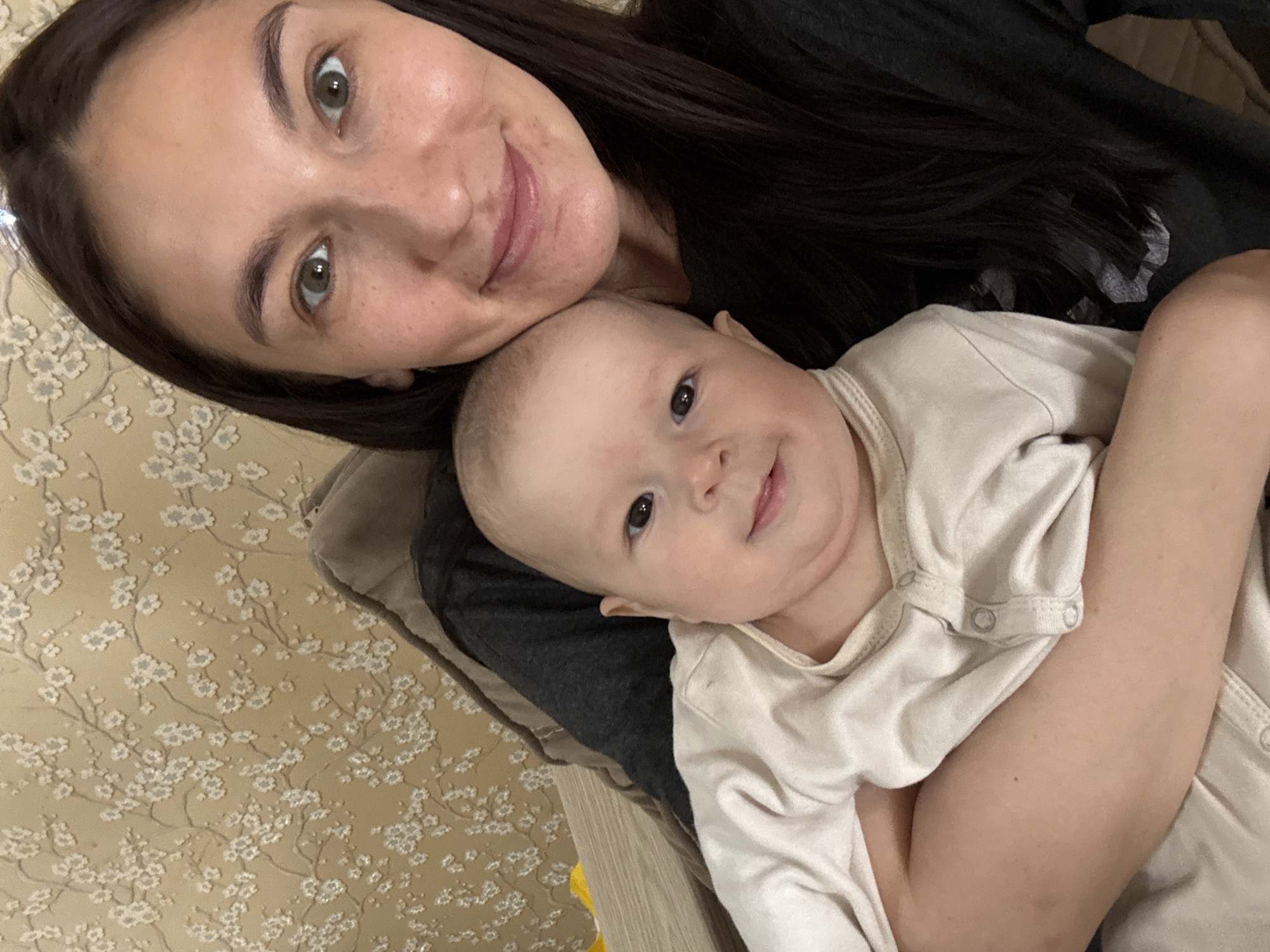 A woman with dark hair and fair skin is taking a selfie with a baby. The baby is wearing a white onesie and is lying on the woman's chest. The woman is smiling at the camera. The background appears to be an indoor scene with patterned wallpaper. There's no information to support any assumptions about a location in the city of Sochi, Russia.