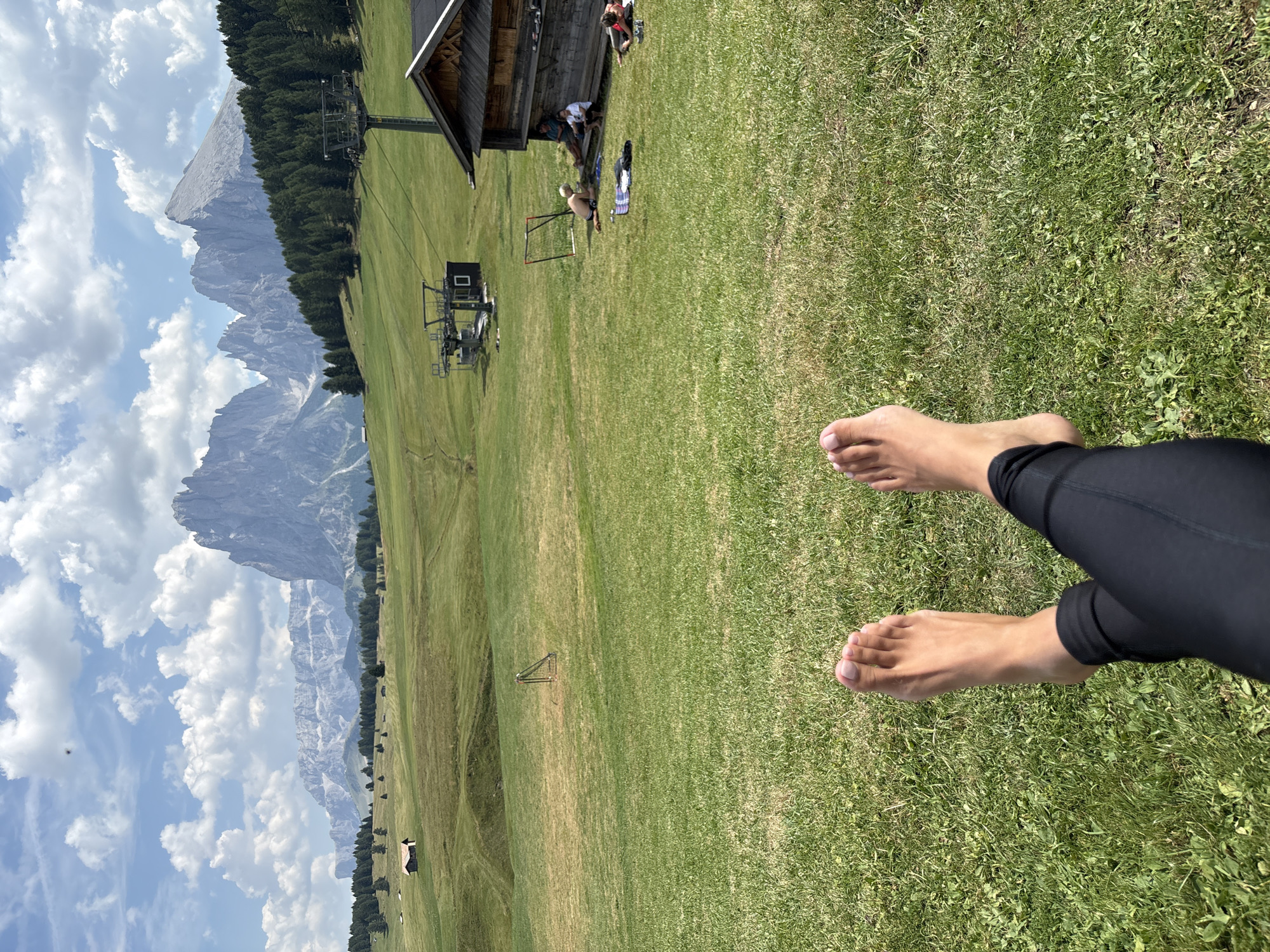 The image captures a scenic view of the Dolomite mountains, likely from the Alpe di Siusi (Seiser Alm) in Kastelruth - Castelrotto, Italy. In the foreground, the bare feet and lower legs of a person wearing black leggings are visible, positioned on a grassy slope. The mid-ground features rolling green hills, dotted with scattered trees and a few small buildings. A ski lift station with a tower and cable car structure is prominently situated on the right side of the frame, adjacent to a rustic wooden building. Several people are seen relaxing in the vicinity of the building, some sitting on the ground and others near the wooden structure. The background is dominated by the majestic, jagged peaks of the Dolomites, illuminated by bright sunlight under a partly cloudy sky.