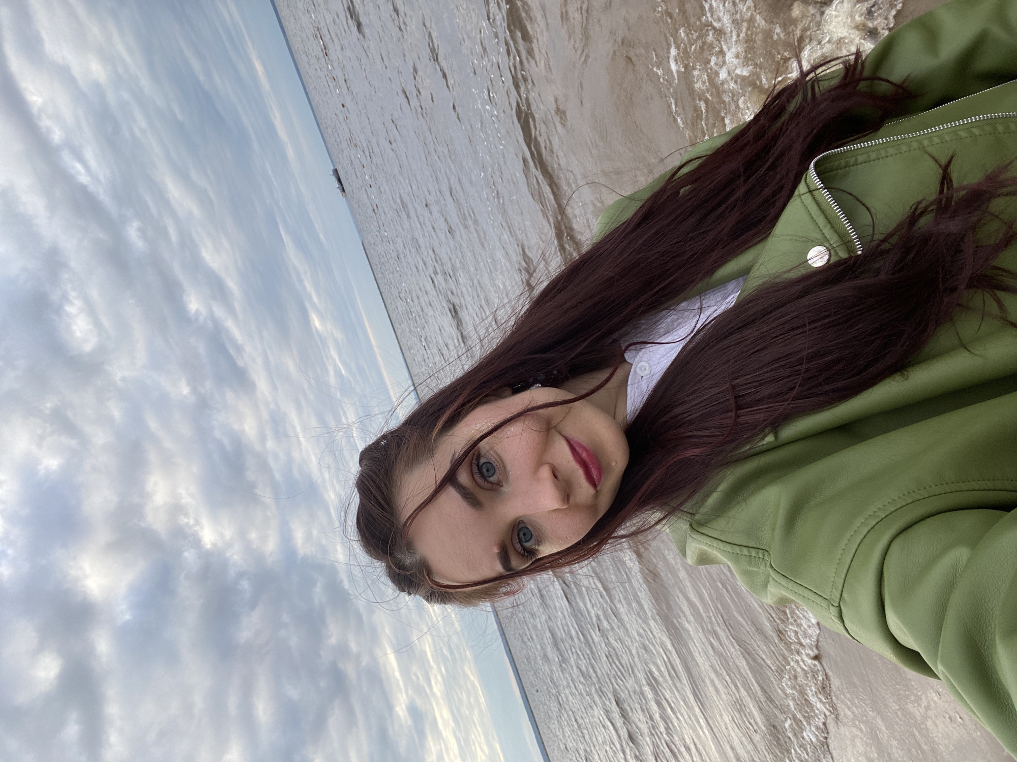 A young woman with long, dark red hair and striking blue eyes is captured in a selfie, taken outdoors. She is wearing a white shirt and a green jacket. Her expression is a slight smile, and she has applied dark pink lipstick. Her hair is partly covering her face, and some strands are being blown by the wind. The background is a body of water, likely a river or sea, with a cloudy sky above. There is a faint outline of a ship on the horizon. The lighting suggests it is daytime, possibly late afternoon or early evening, given the soft, diffused light. The presence of a ship on the horizon and the open water suggest a coastal or riverside location. The overall mood of the image is casual and relaxed, hinting at a moment of personal reflection or enjoyment of the natural surroundings.