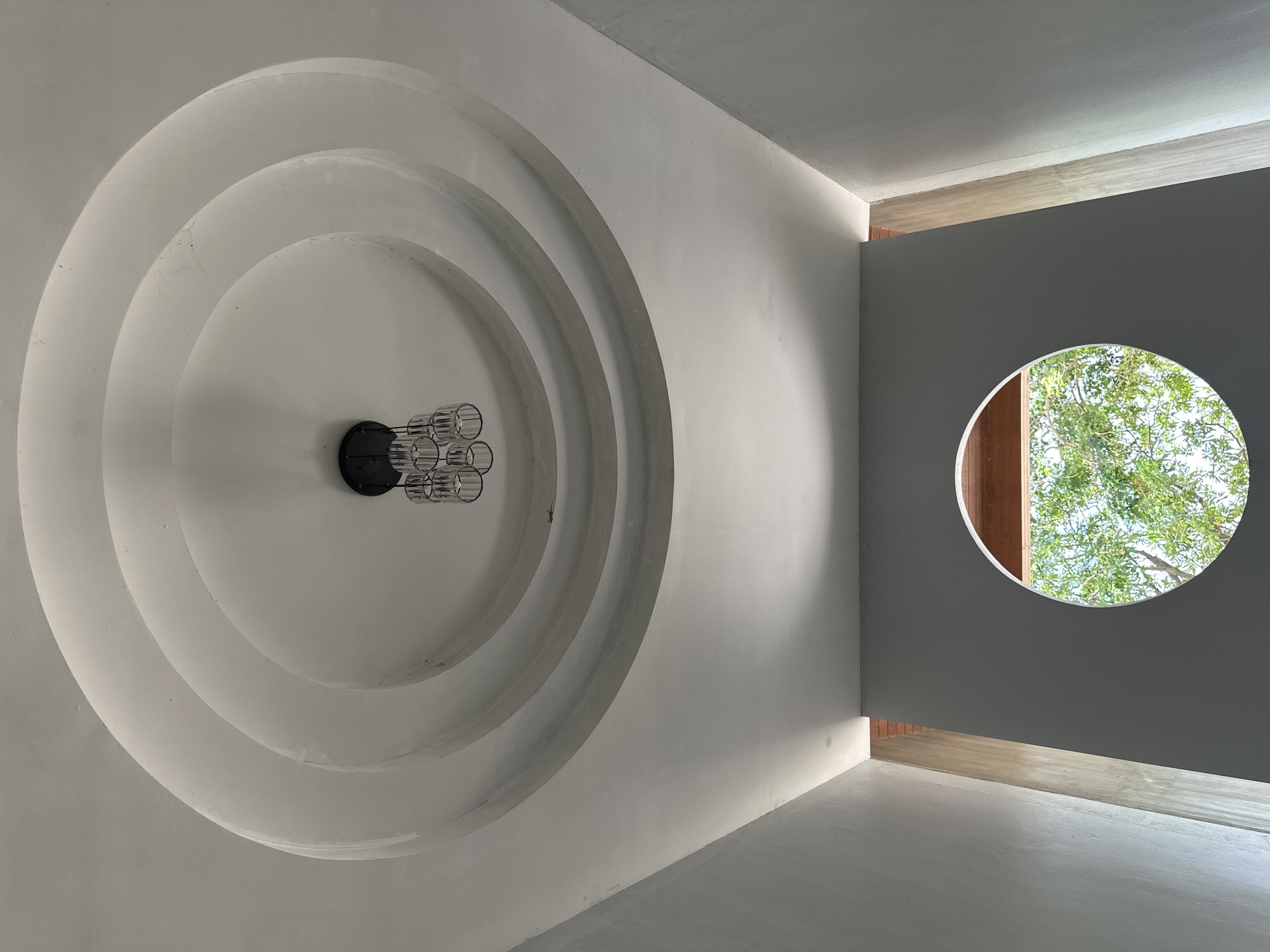 The image depicts the interior of a modern building in ตำบลเกาะพะงัน, Thailand. The foreground shows a ceiling with a decorative, multi-layered circular design, from which a contemporary chandelier with multiple cylindrical glass shades hangs. Below the ceiling, a circular window is visible, offering a view of lush green trees and the sky. The walls are painted in neutral tones, with a textured finish on the left side, suggesting a contemporary architectural style. The scene is devoid of people or any visible activity, focusing solely on the architectural elements and the natural view through the window.