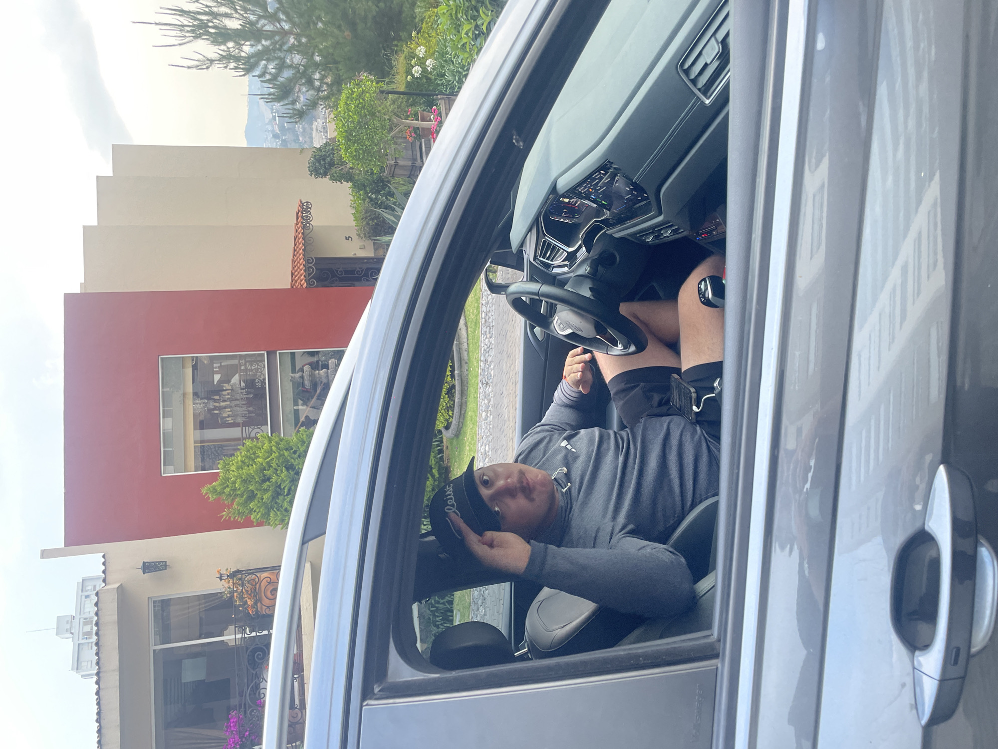 A man is sitting in the driver's seat of a gray vehicle. He is wearing a gray long-sleeved shirt, black shorts with compression leggings underneath, and a black baseball cap with the word "Titleist" on the front. He appears to be looking at the camera and adjusting his cap. The dashboard and steering wheel of the car are visible. Through the open car door, a building with a red and beige exterior is seen, along with greenery. The location is implied to be in Huixquilucan de Degollado, Mexico.