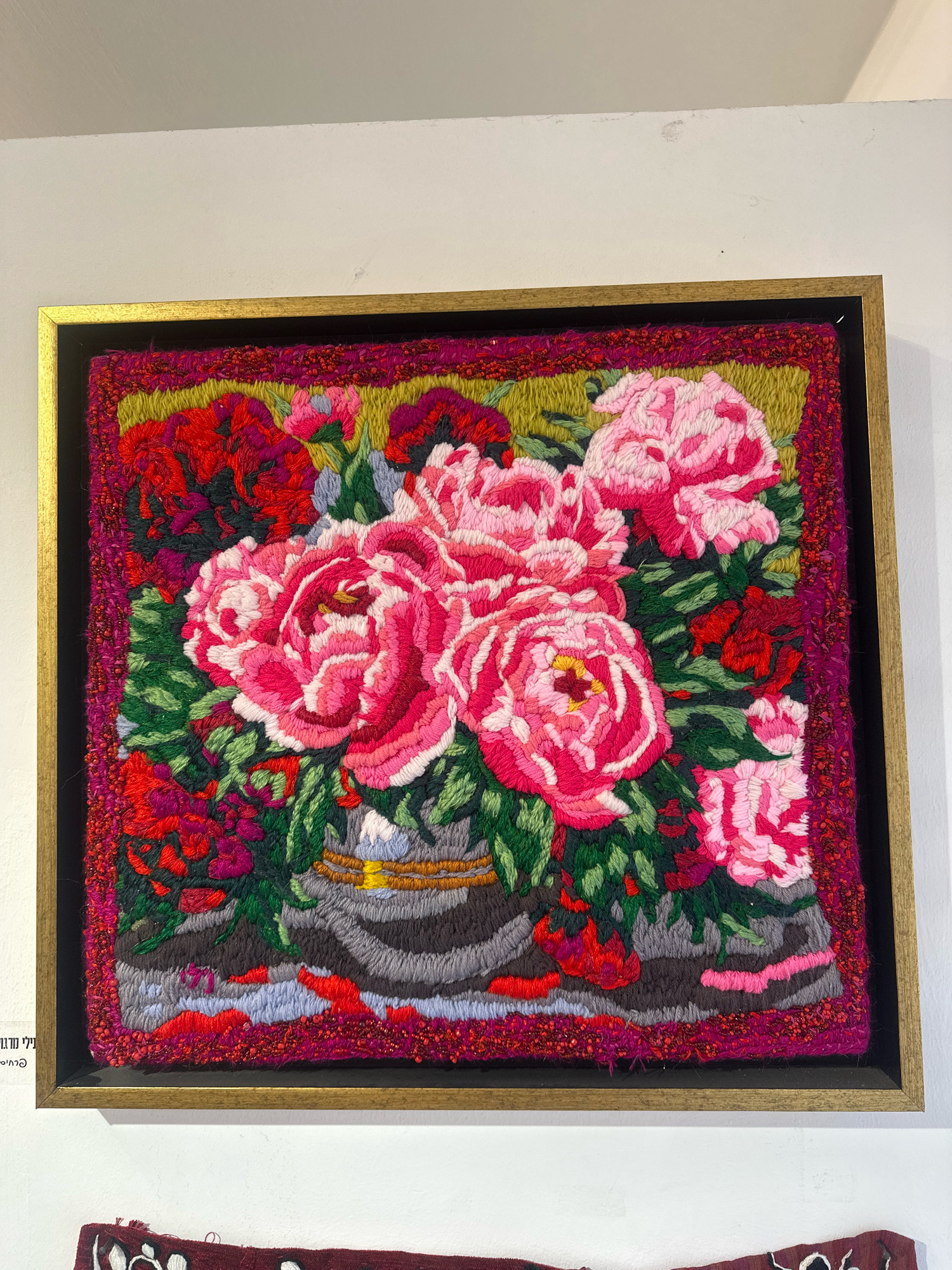 This is a close-up, eye-level shot of a framed textile art piece, possibly a tapestry or rug, depicting a vibrant floral arrangement. The artwork is hanging on a plain white wall, likely in an exhibition or gallery space. The dominant colors are various shades of pink, red, and green, with accents of yellow, blue, and grey. The flowers, primarily large, textured blossoms resembling peonies, are centrally located. They are arranged in a vase, the base of which is visible at the bottom of the artwork. The background of the artwork is a textured, dark green. The framing consists of a black matting around the textile, surrounded by a textured, magenta border with small red beads. This is further encased by a thin, golden frame. To the left of the artwork, a small portion of text in Hebrew script is visible, suggesting a label or inscription. Below the framed artwork, the corner of another textile piece with white embroidery on a dark red background is visible. The overall lighting is soft and diffused, typical of indoor exhibition spaces. The scene is located in Mate Asher Regional Council, Israel, as indicated by the Hebrew text on the label.