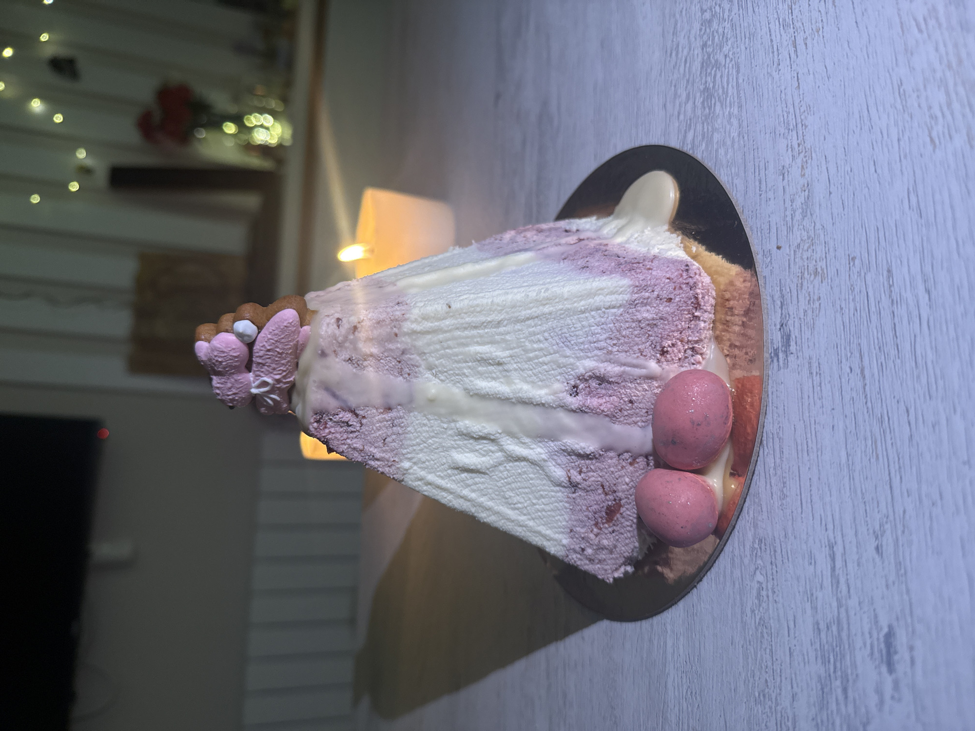 A slice of layered cake rests on a gold-colored plate. The cake has pink and white layers with white frosting drizzled over the top. It is decorated with two pink eggs and a cluster of pink and brown decorations. The background is blurred, but includes a candle, and some indistinct shapes that might be part of a room. The location is indeterminable.