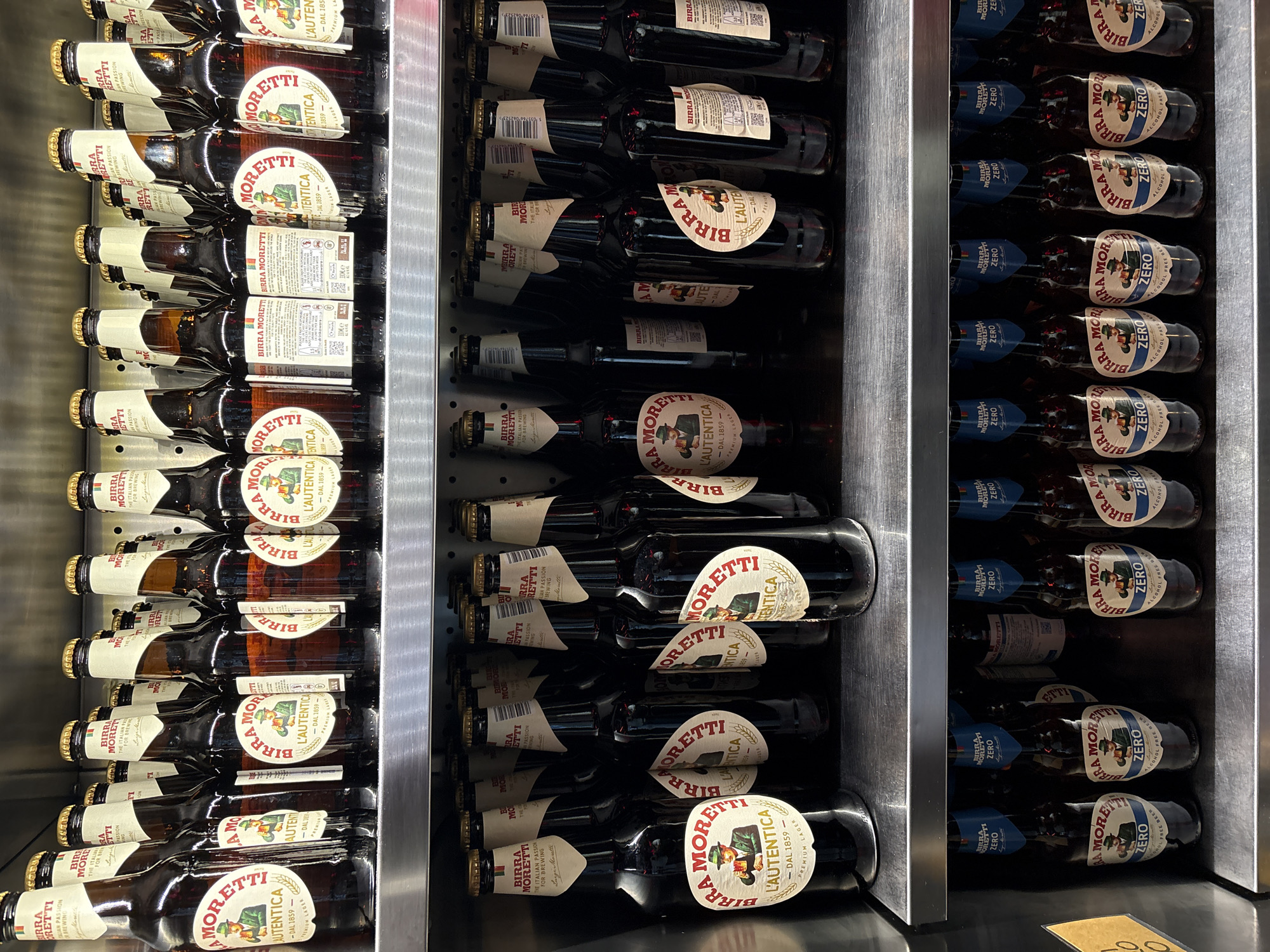 The image shows a refrigerated display case containing three rows of Birra Moretti beer bottles. The left row contains Birra Moretti L'Autentica, the middle row contains a darker variant of the same beer, and the right row contains Birra Moretti Zero alcohol-free beer. The bottles are neatly arranged horizontally within the metal-framed display case. The background is out of focus. There are no people visible.