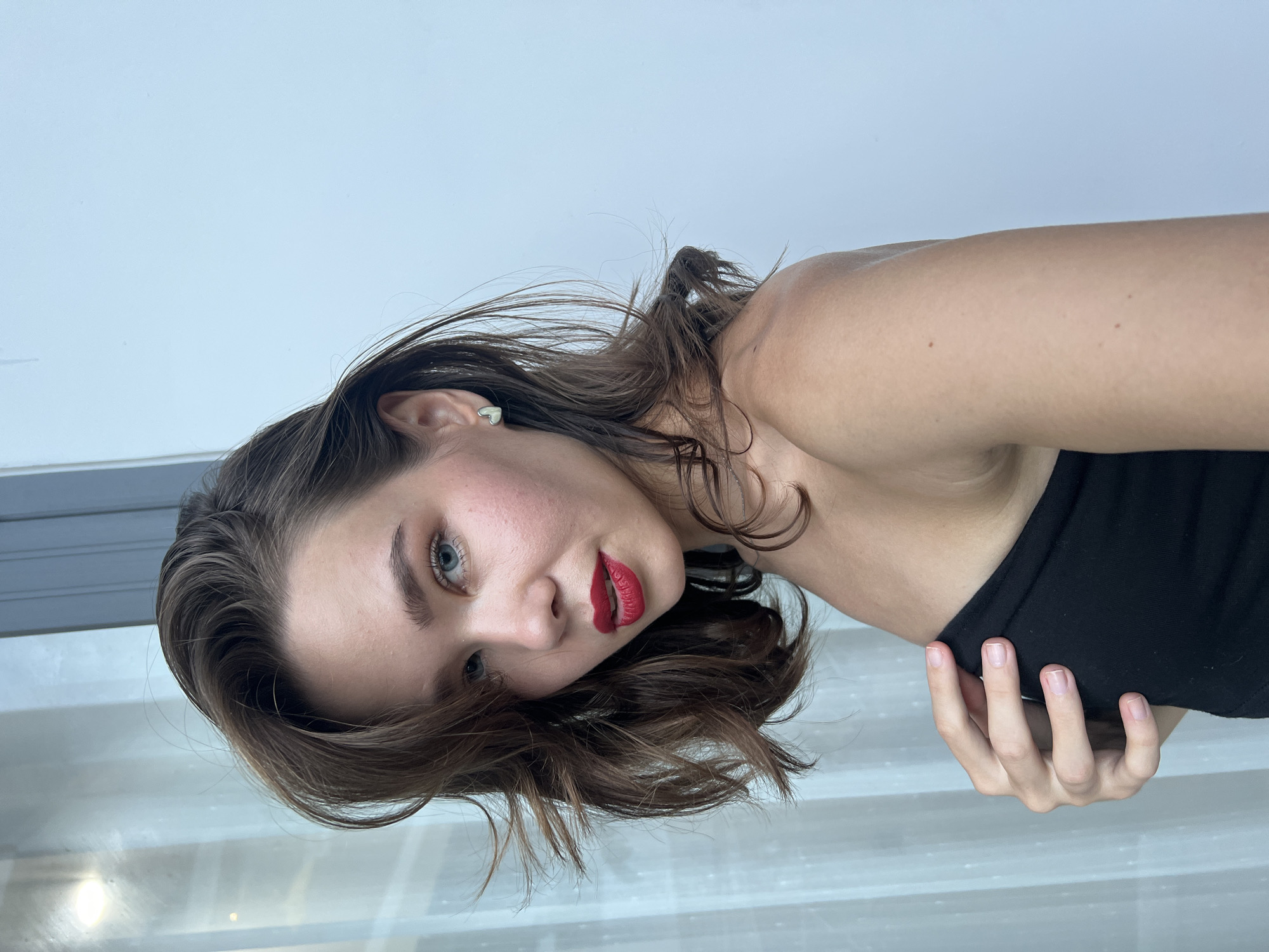 A woman with fair skin, red lipstick, and brown hair is looking at the camera. She is wearing a black top. She is indoors with white walls and what appears to be a window or reflective surface below her. There is no visible evidence to confirm the location within Nam Nha Trang Ward, Vietnam.