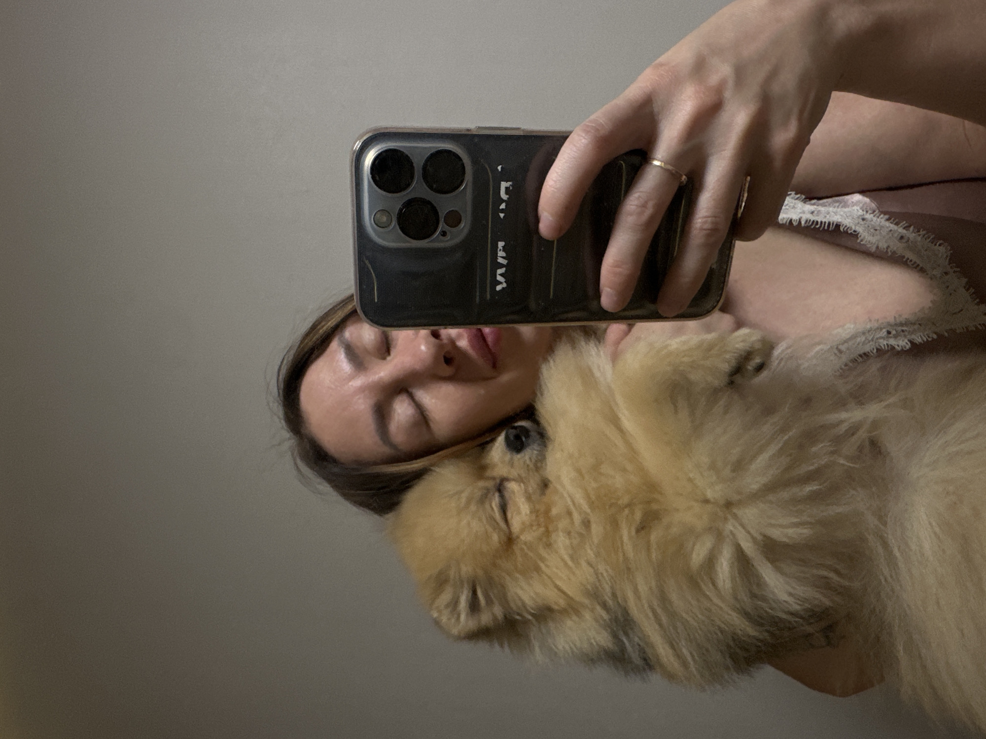 In this close-up, waist-up, selfie-style photograph, a woman with brown hair and closed eyes is holding a small, fluffy, tan-colored Pomeranian dog. The dog is nestled against the woman's chest and appears to be asleep, with its eyes closed. The woman is holding a dark-colored smartphone in her right hand, angled to capture the reflection and the dog. She is wearing a thin-strapped, pink garment with white lace trim. A thin gold ring is visible on her left ring finger. The background is a plain, muted gray wall, suggesting an indoor setting. The lighting is soft and diffused. The image does not provide any specific geographic or environmental clues that would allow for determination of the location within Votkinsk, Russia.