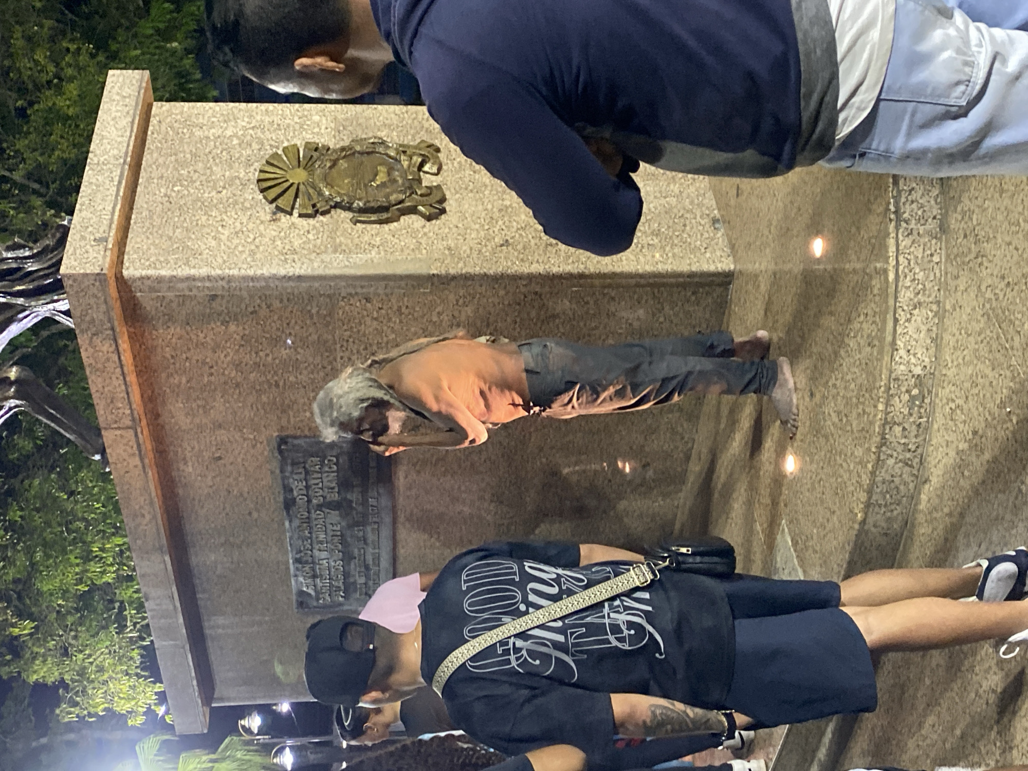 In Tinaquillo, Venezuela, a man with long, gray hair and no shirt stands near a monument. He is wearing dark pants and is barefoot. He appears to be touching his face. To his left, a person wearing a black t-shirt with "GOOD THINGS" written on it and a crossbody bag stands facing away from the camera. To the right of the monument, another person wearing a dark blue hoodie and light blue pants is partially visible, facing away from the camera. The monument is made of speckled stone and has a plaque on it with text that is difficult to read. A decorative emblem is also affixed to the monument. The environment appears to be outdoors, with trees and streetlights visible in the background, suggesting it is nighttime. There are small lights on the ground around the monument.