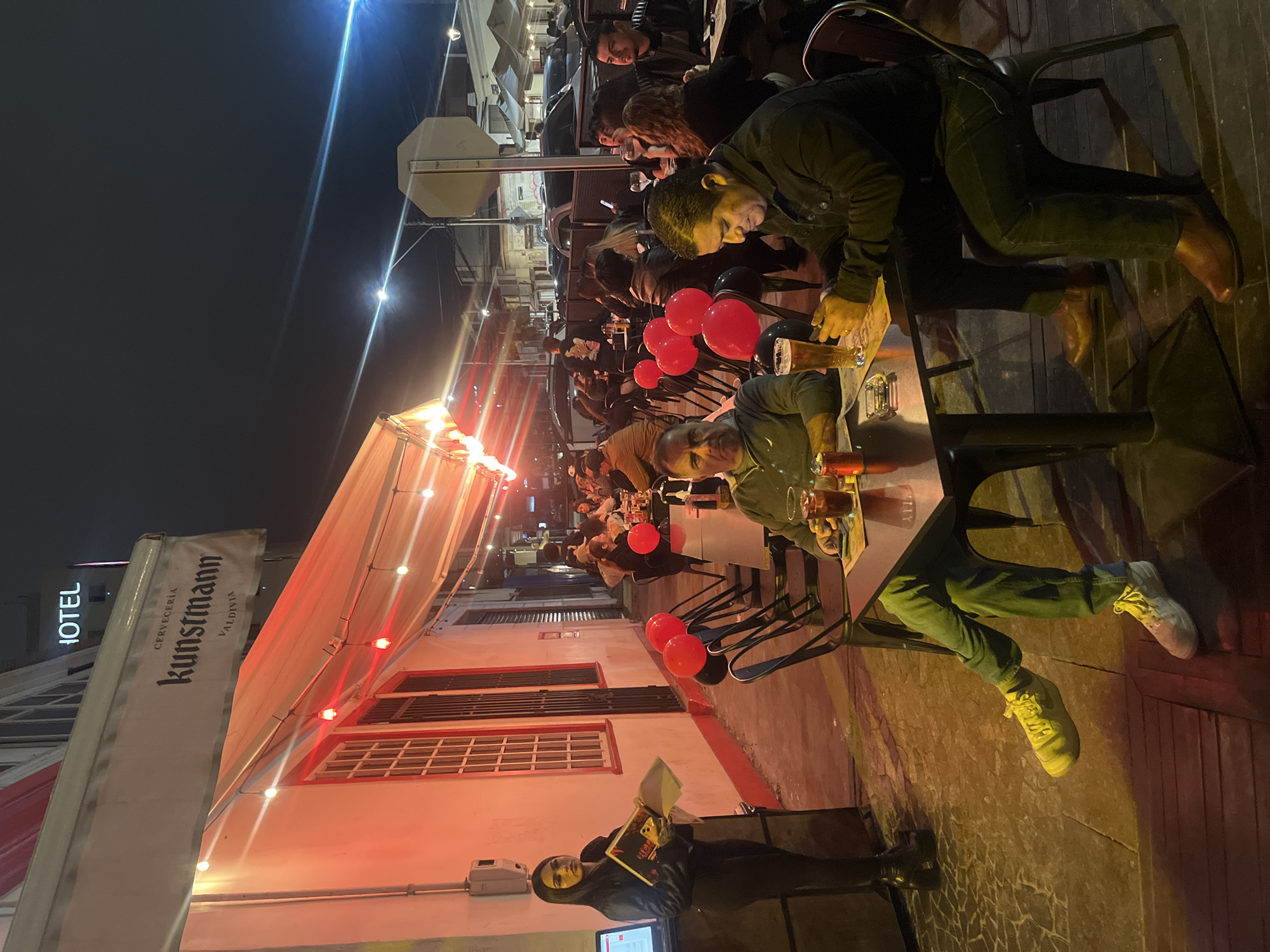 An outdoor seating area of a restaurant in Iquique, Chile, is depicted at night. A woman in a black jacket and pants stands near a counter with a computer screen, holding papers. Several tables with patrons are visible, some with red and black balloons attached to chairs. People are seated at the tables, some with drinks and food. The restaurant has a white awning with red lights strung underneath. The overall atmosphere appears to be a casual dining experience at night.