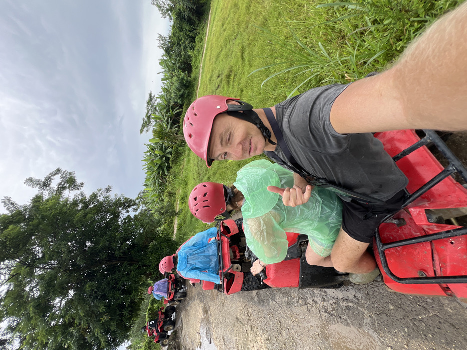 A selfie of a group on red ATVs, stopped on a muddy path. The first two figures are wearing pink helmets; the rest are obscured by rain ponchos in green, blue, and purple. The foremost person, a man, wears a gray shirt and gives a slight smile. The second person, also in a pink helmet, wears a green poncho and holds up their index finger. Lush green foliage and a cloudy sky form the backdrop.