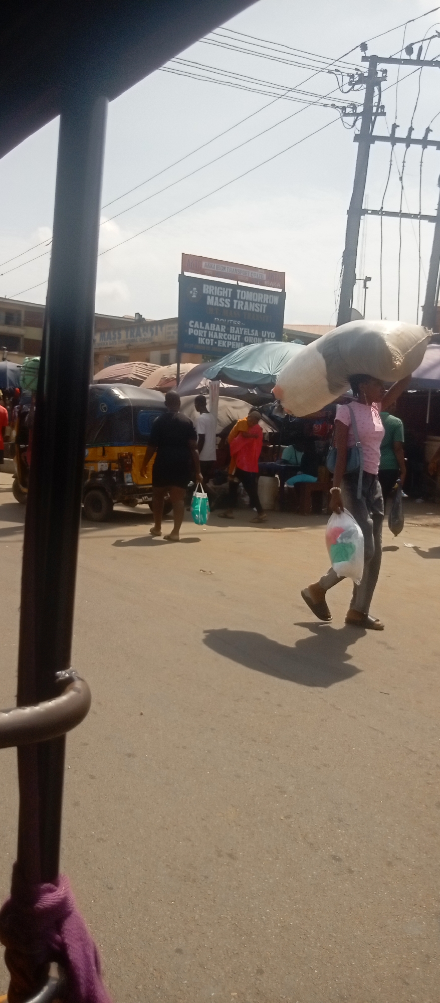 The image captures a street scene in Onitsha, Nigeria, bustling with activity. The focus is on a woman walking along the road, carrying a large sack on her head and a plastic bag in her hand. Other pedestrians are visible in the background, along with a yellow tricycle (keke) and market stalls covered with tarpaulins. A sign for "Bright Tomorrow Mass Transit" is prominently displayed, advertising routes to Calabar, Bayelsa, Uyo, Port Harcourt, Oron, and Ikot-Ekpene. Overhead, utility poles and wires crisscross the sky. The scene suggests a typical busy day in a Nigerian city. The viewpoint is from inside a vehicle.