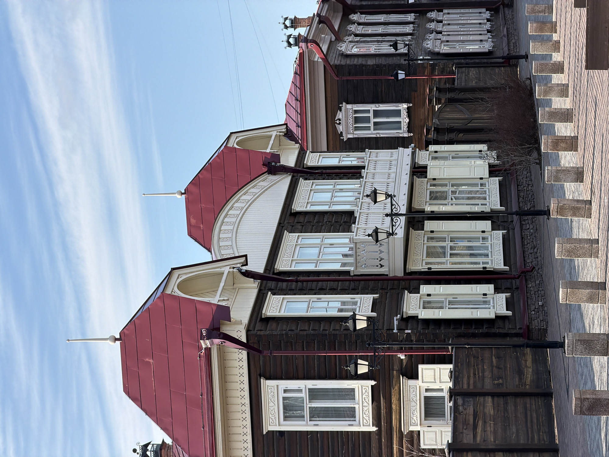 A vertically oriented, medium shot captures a row of ornate wooden buildings in Krasnoyarsk, Russia, under a blue sky with streaks of clouds. The buildings feature dark-stained wood siding contrasted by elaborate white trim around the windows and along the eaves. The roofs are painted in a shade of red. Victorian-style street lamps are positioned in front of the buildings. A paved walkway runs parallel to the buildings, marked by small, square stone blocks. The scene has a clear, bright daylight setting.
