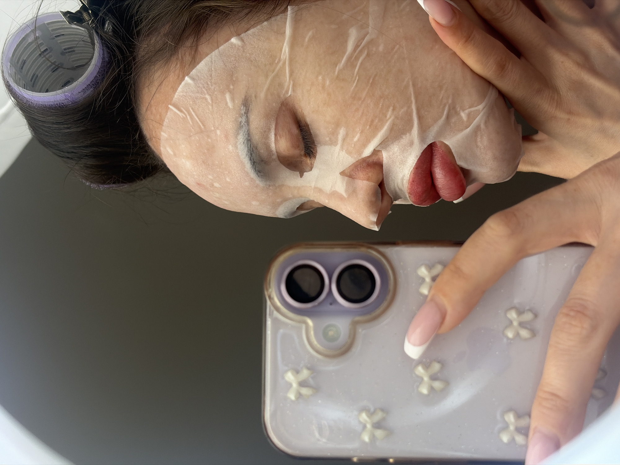 A woman wearing a sheet face mask and hair rollers is taking a selfie. The phone has a lavender case decorated with white clover shapes. The woman has manicured nails with a white French tip. The background is a blurred gray. There are no indications of Saint Petersburg, Russia in the image.