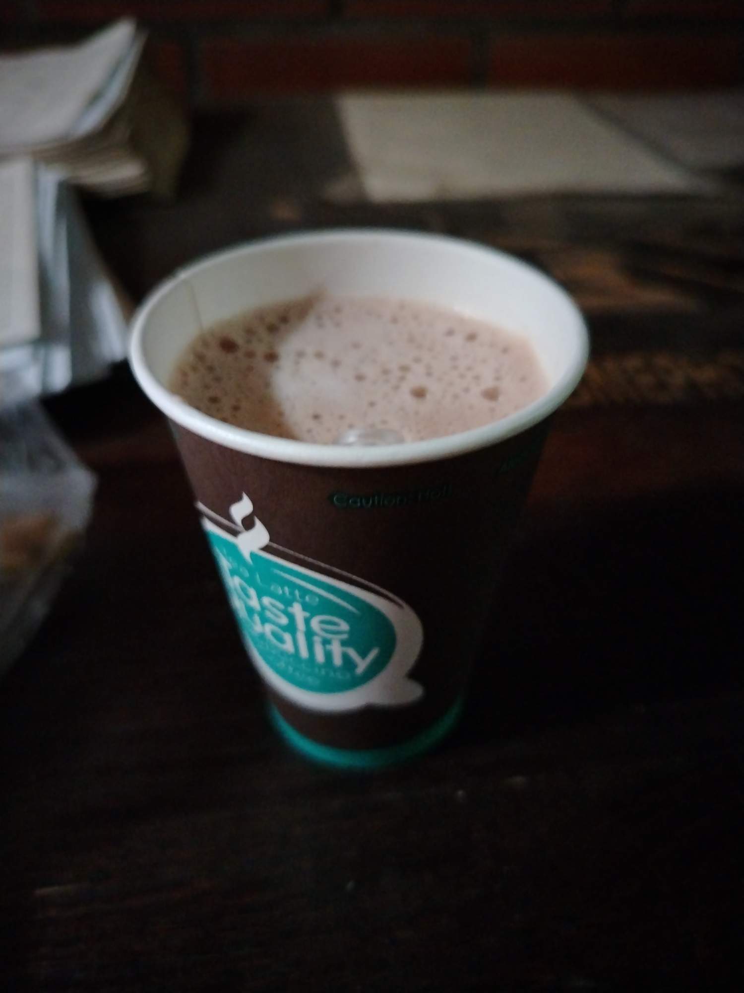 A close-up, slightly blurry image captures a brown paper cup containing a frothy, light-brown beverage, likely hot chocolate or coffee, on a dark wooden surface. The liquid exhibits numerous small bubbles on its surface, indicating it is a warm or hot drink. The cup itself is brown with a distinctive teal and white label design. Prominently displayed in white, cursive-style lettering on the teal background are the words "taste quality" with "latte" written above it. Just above this main label, a faint warning text, "Caution Hot," is visible on the brown part of the cup.
The background is largely out of focus, showing indistinct shapes and colors. To the upper left, there appears to be a stack of papers or documents. Behind these, a reddish-brown, brick-like pattern can be vaguely discerned, suggesting an indoor setting with a brick wall or similar architectural feature. The overall lighting is somewhat dim, highlighting the cup and its contents. No people or specific activities are shown, but the presence of the beverage implies it is prepared for consumption. The scene is set in an unspecified indoor location, with the provided context indicating it is in Шебунино, Russia.