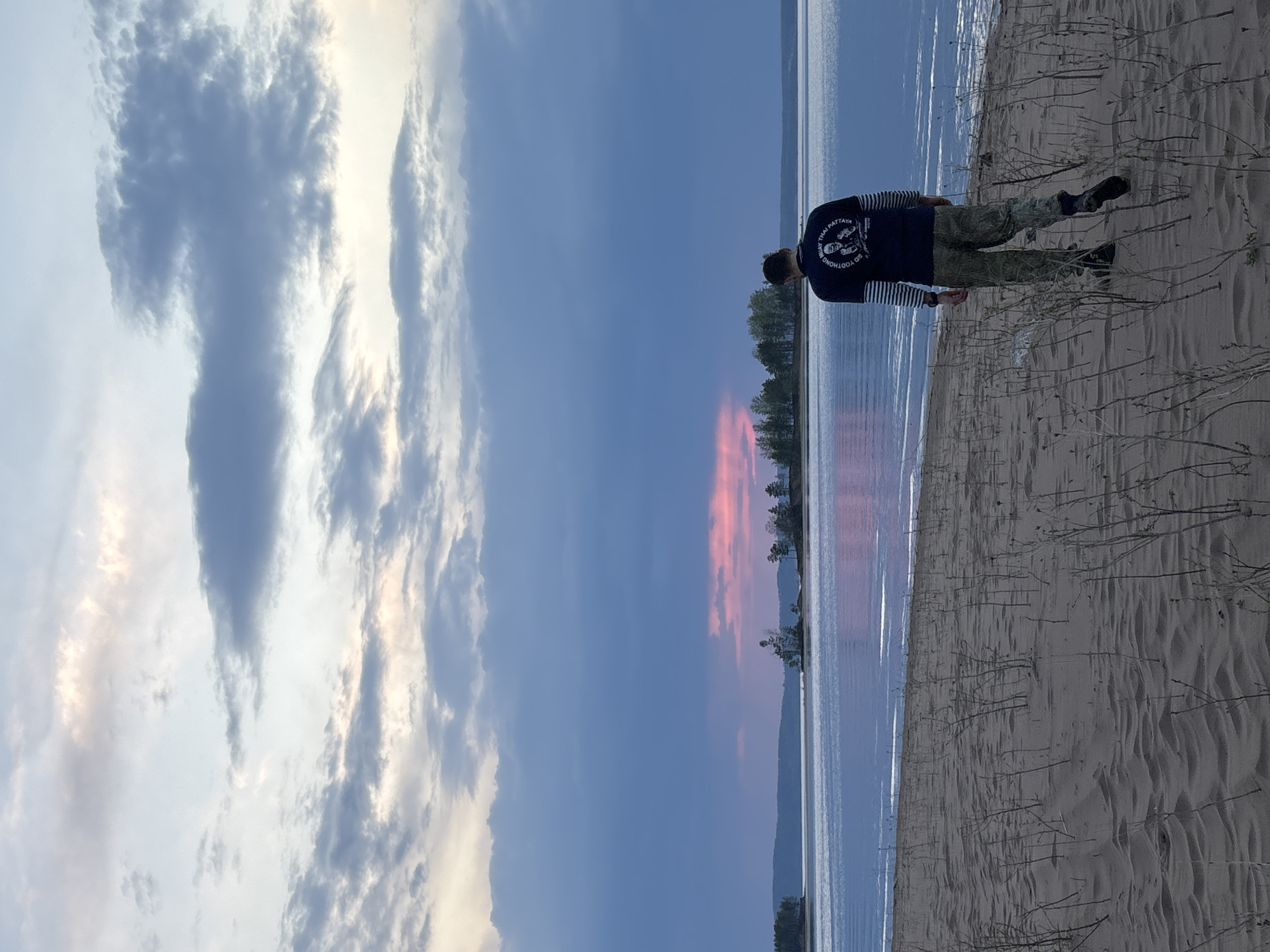 The image shows a man standing on a sandy beach in Vekoshinka, Russia. The man is wearing a dark blue shirt with white horizontal stripes on the sleeves and camouflage pants. He is facing away from the camera, looking out towards a body of water that appears to be a lake or river. The sky is a mix of blue and pink hues, suggesting either a sunrise or sunset. Clouds are visible in the sky. The foreground is sandy with some small vegetation.