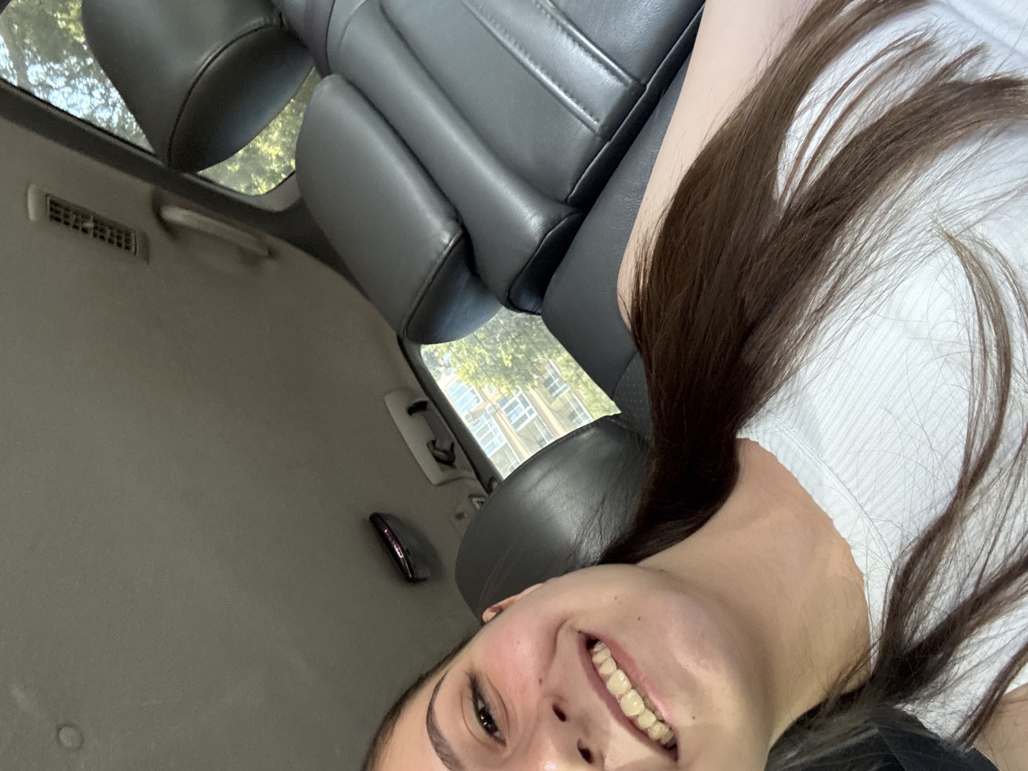 The image is a brightly lit, vertical selfie taken inside a car in Burgas, Bulgaria. A young woman, likely in her late teens or twenties, is the primary subject, occupying the lower right portion of the frame. She has long, dark brown hair that falls over her shoulders and a white, ribbed top. Her face is turned towards the camera, and she is smiling broadly with her mouth open, revealing her teeth, conveying a happy and relaxed emotion. She has dark eyebrows and dark eyes.
The setting is the interior of a car, seen from a low angle, suggesting the woman is a passenger, possibly in the back seat. Dark gray or black leather car seats are visible behind her, including headrests and seatbacks. The car's ceiling, or headliner, is a light gray or tan color, featuring an air vent and a grab handle. A dark, oblong object, possibly a clip or holder, is also attached to the ceiling towards the top left.
Through the car windows, lush green foliage from trees is visible, indicating an outdoor, daytime setting. In the background, beyond the trees, a light-colored building with multiple windows can be seen. The bright lighting suggests it is daytime, possibly sunny or partly cloudy. There is no visible text in the image.