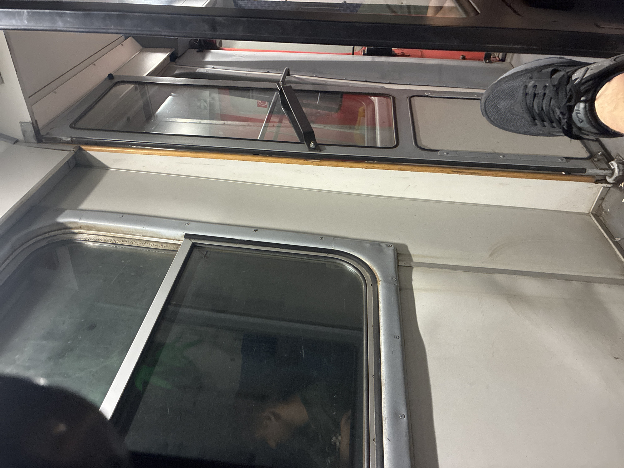 The image shows the interior of a train car, possibly a subway or a regional train, with a focus on the window and the door. A person's foot in a dark sneaker is visible in the lower right foreground, resting on what appears to be the floor of the train. To the left, a window reflects a person's head and shoulders, seen in profile, suggesting they are looking out or resting. The interior of the train car is utilitarian, with light-colored walls and a metal frame around the window. A dark metal handle is attached to the door, which is partially open, revealing red elements inside the train car beyond. The environment appears to be dimly lit, typical of a train's interior, especially if it is in transit or at a station at night. There is no visible indication of the specific location being Skobelevo, Bulgaria, and no details in the image directly support this assertion.