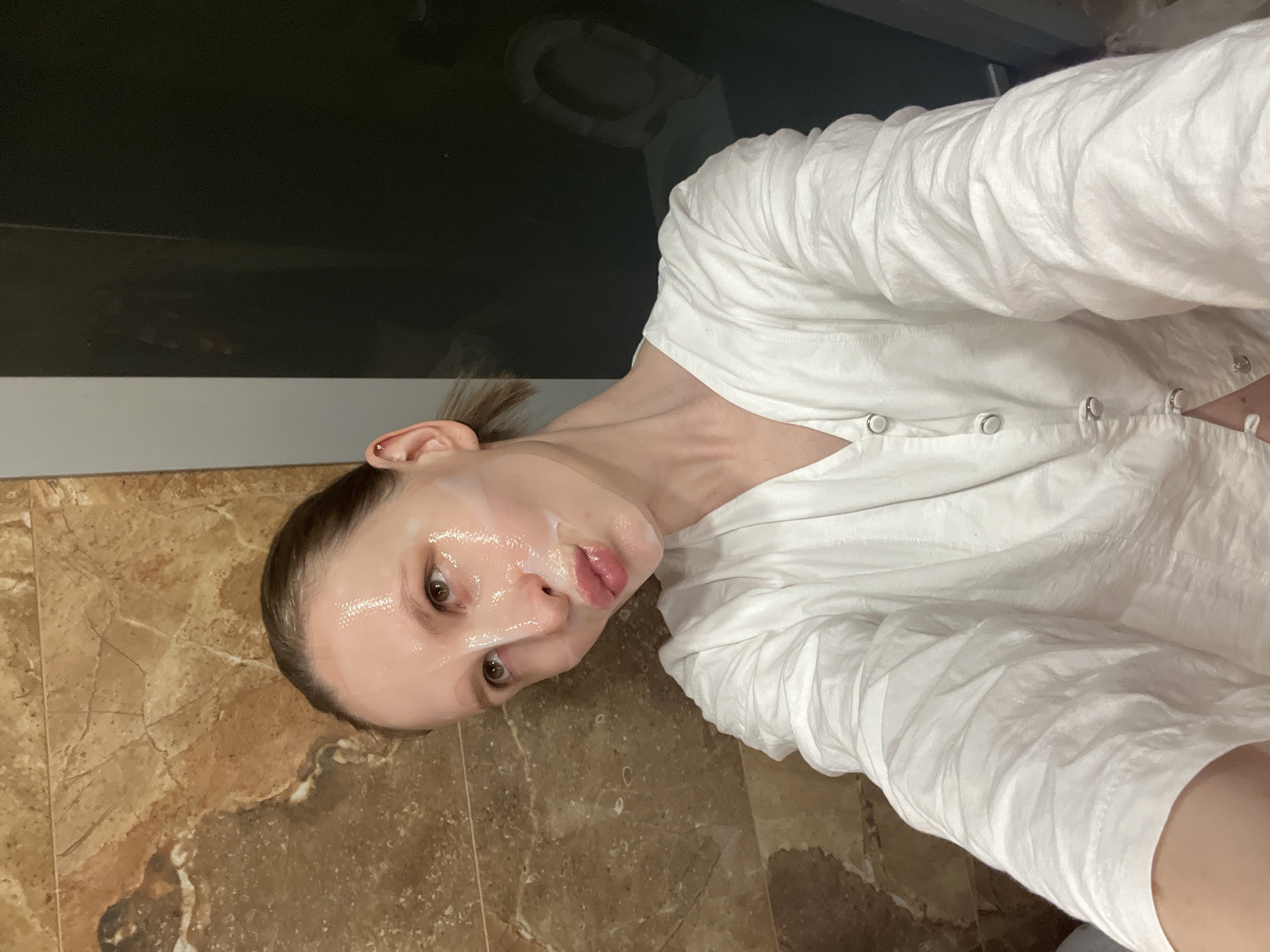 A woman with a face mask lies on a tile floor in what appears to be a bathroom. She is wearing a white, buttoned shirt. The tile floor is brown and marbled. In the background, there is a black wall above the tile. The location is Unknown, Unknown.