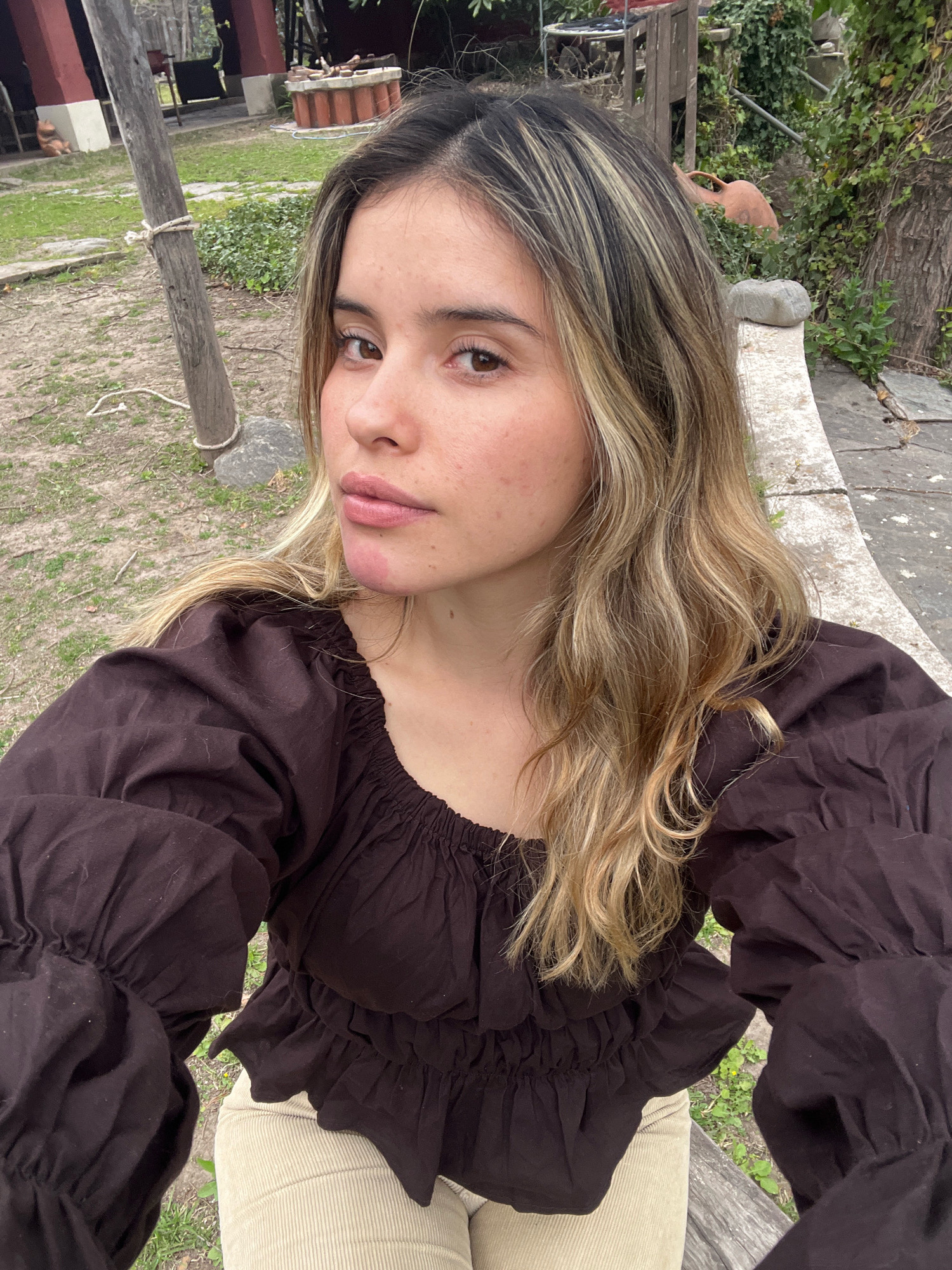 The image shows a young woman with blonde-streaked brown hair posing for a selfie outdoors. She is wearing a dark brown puff-sleeved blouse and light-colored corduroy pants. The background suggests a semi-rural or garden setting, with grass, trees, and some outdoor structures visible. Specifically, the location is identified as El Rodeo, Argentina. A wooden post wrapped with rope is prominent in the left foreground.