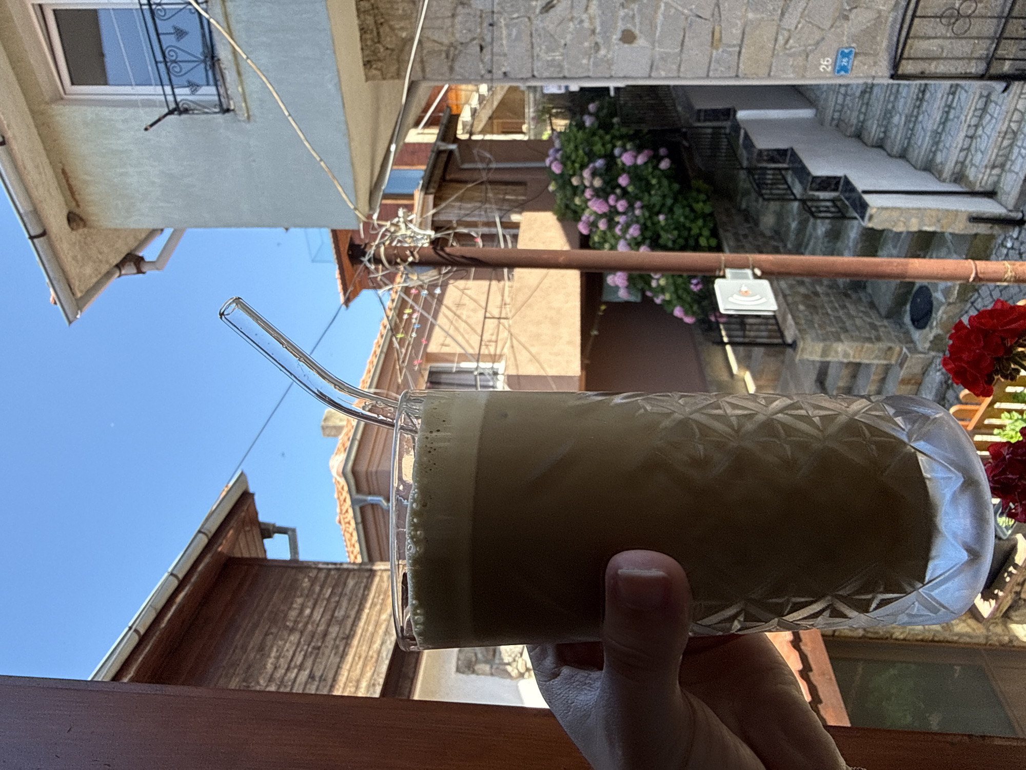 A hand holds a clear glass mug filled with a frothy, light brown beverage. The mug has a patterned texture around its base. A clear glass straw is placed in the drink. The background shows a sunlit outdoor scene with buildings and a clear blue sky. The buildings appear to be of stone and stucco construction with terracotta tile roofs. In the mid-ground, a bush with pink flowers is visible, and a set of stone stairs ascends between buildings. A blue sign with white text and a number "26" is affixed to a stone wall near the top of the stairs. A metal railing runs alongside the stairs. In the foreground, the image is partially framed by wooden surfaces. The overall impression is a bright, sunny day in a European town.