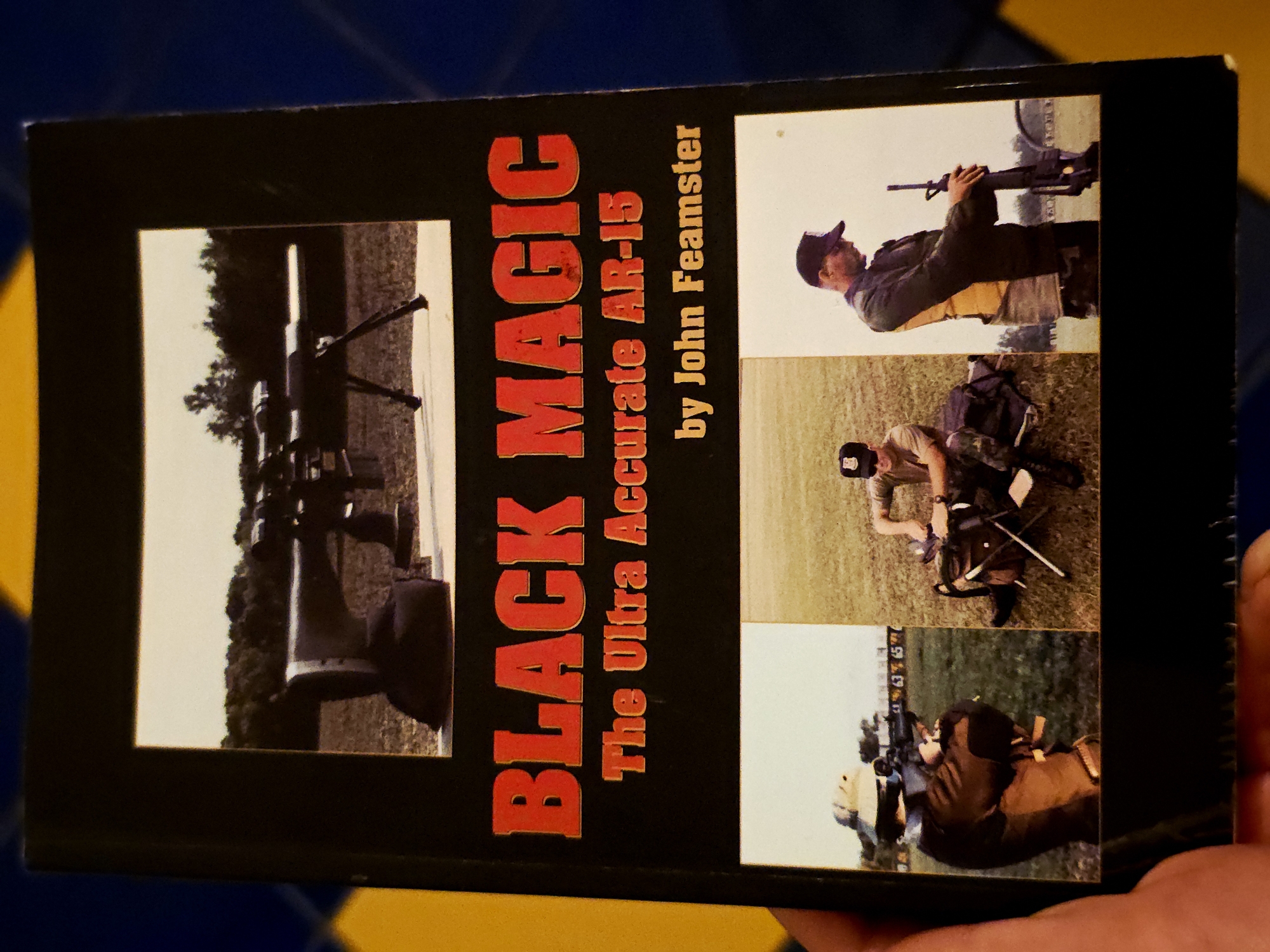 This is an image of the front cover of a book titled "BLACK MAGIC The Ultra Accurate AR-15 by John Feamster." The cover features a black background with bold red and orange/gold text.
The title "BLACK MAGIC" is prominently displayed in large red letters running vertically down the left side of the cover. Below this, in smaller orange/gold text, is "The Ultra Accurate AR-15," followed by "by John Feamster."
The right side of the cover contains three individual photographs, arranged vertically, depicting men engaged in rifle-related activities in an outdoor setting.
1. The top photo shows a man in a dark cap and jacket holding an AR-15 style rifle, looking forward.
2. The middle photo features a man in a cap and t-shirt, kneeling on a grassy field, seemingly adjusting equipment or a rifle bag with a spotting scope nearby.
3. The bottom photo shows a man, partially visible, aiming a rifle (likely an AR-15 with a scope) from a prone or kneeling position, with target stands visible in the background.
Additionally, a larger, close-up photograph occupies the lower-left section of the cover, showing an AR-15 rifle equipped with a large scope and bipod, positioned on what appears to be a shooting rest or table on a grassy field. All scenes suggest daylight conditions, likely at a shooting range or outdoor practice area. The overall theme of the book cover revolves around accuracy and the AR-15 rifle.